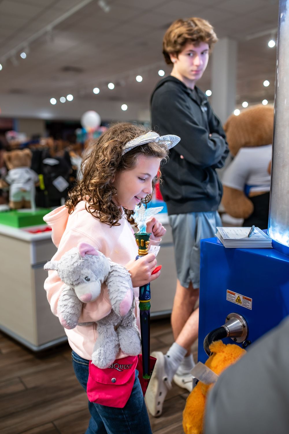 A young child in a white dress holds a plush toy while standing in an indoor space with blue displays.