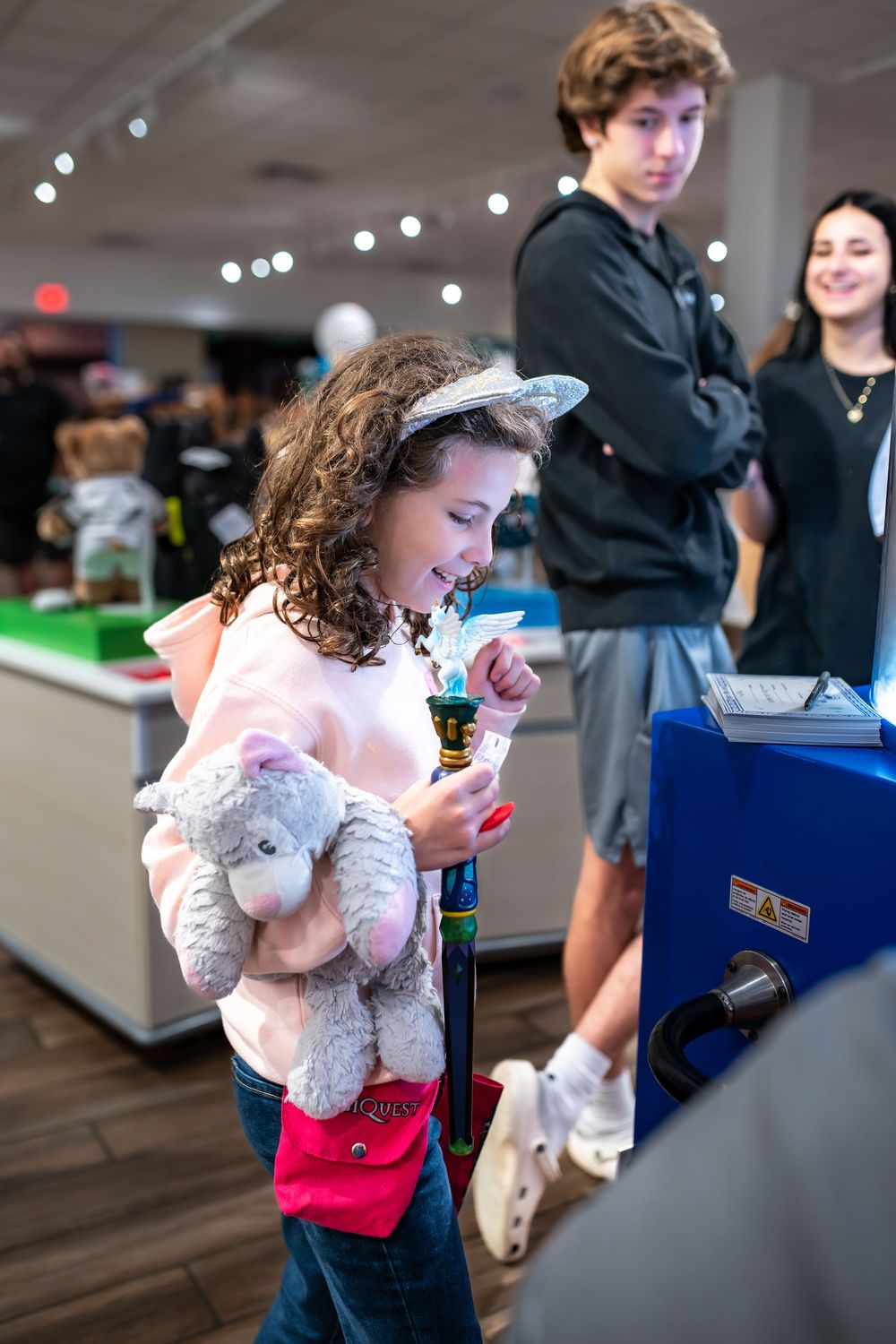 A young child in a white lace dress holds a stuffed animal while looking at a mobile device at an indoor event.
