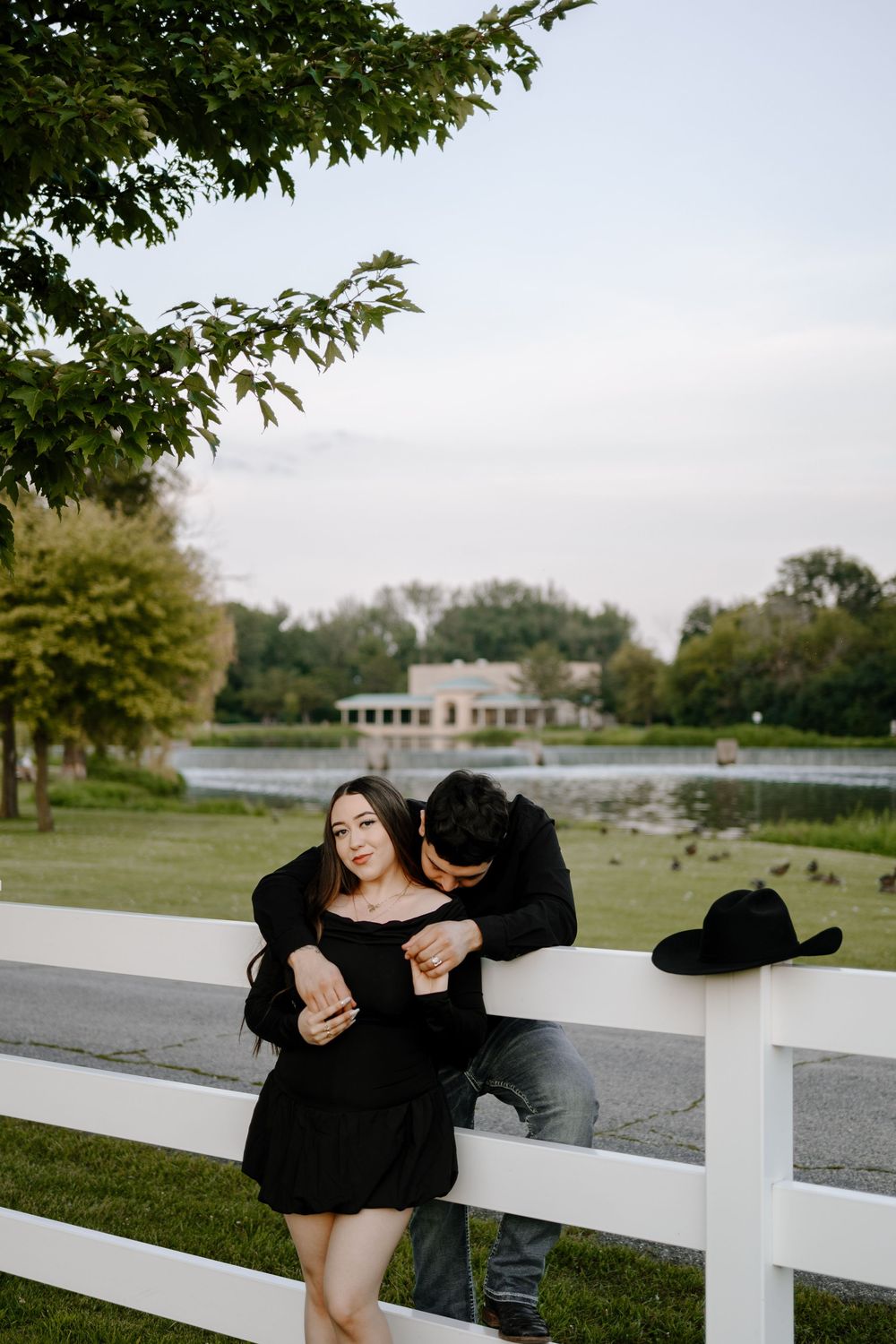 A couple shares a playful moment together while sitting on a white fence near a lake on a sunny day.