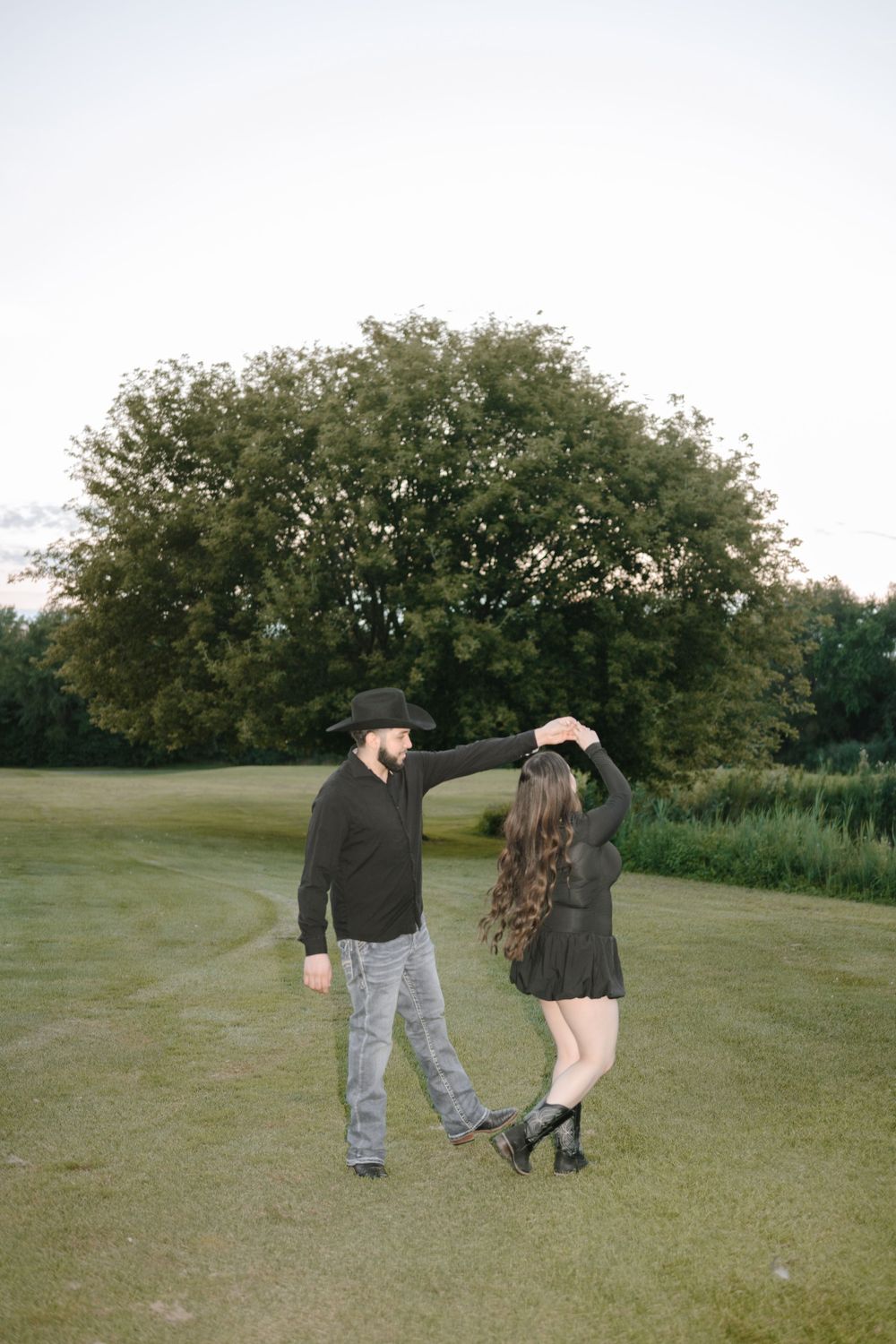 A couple dances playfully in a grassy field with trees in the background.