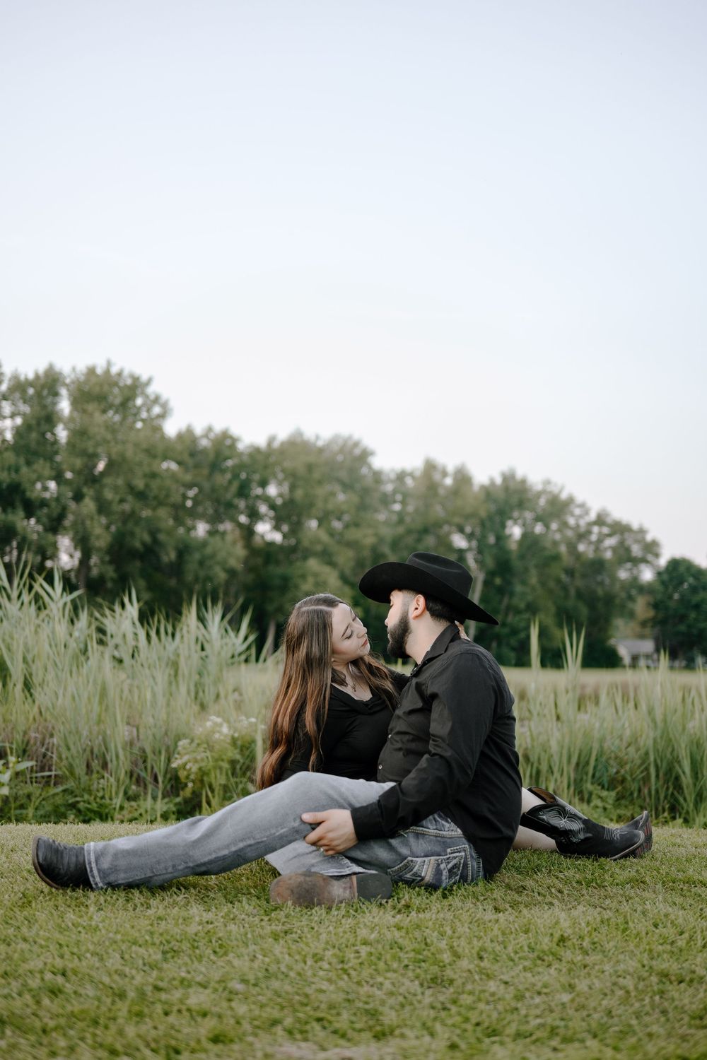 A romantic couple sits together on grass near tall reeds and trees on a peaceful afternoon.