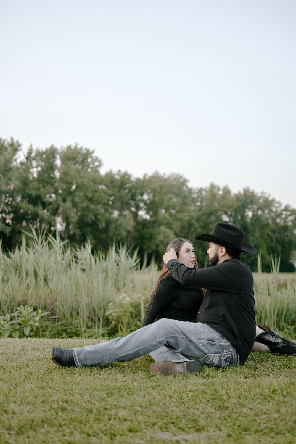 Two people in dark clothing lay together on grass with tall reeds in the background.
