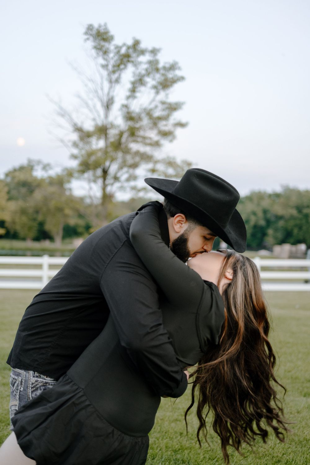 A romantic couple shares an intimate dip pose in a scenic field with white fencing in the background.