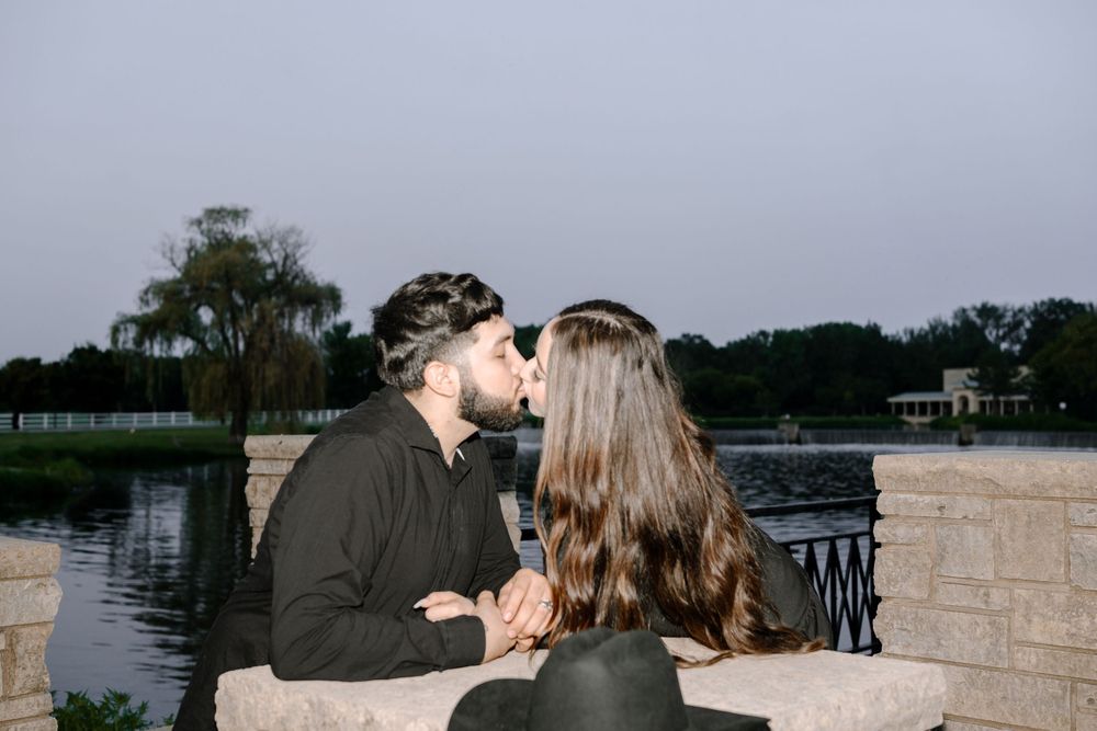 A couple shares a romantic moment at sunset by a stone wall overlooking a scenic waterfront view.