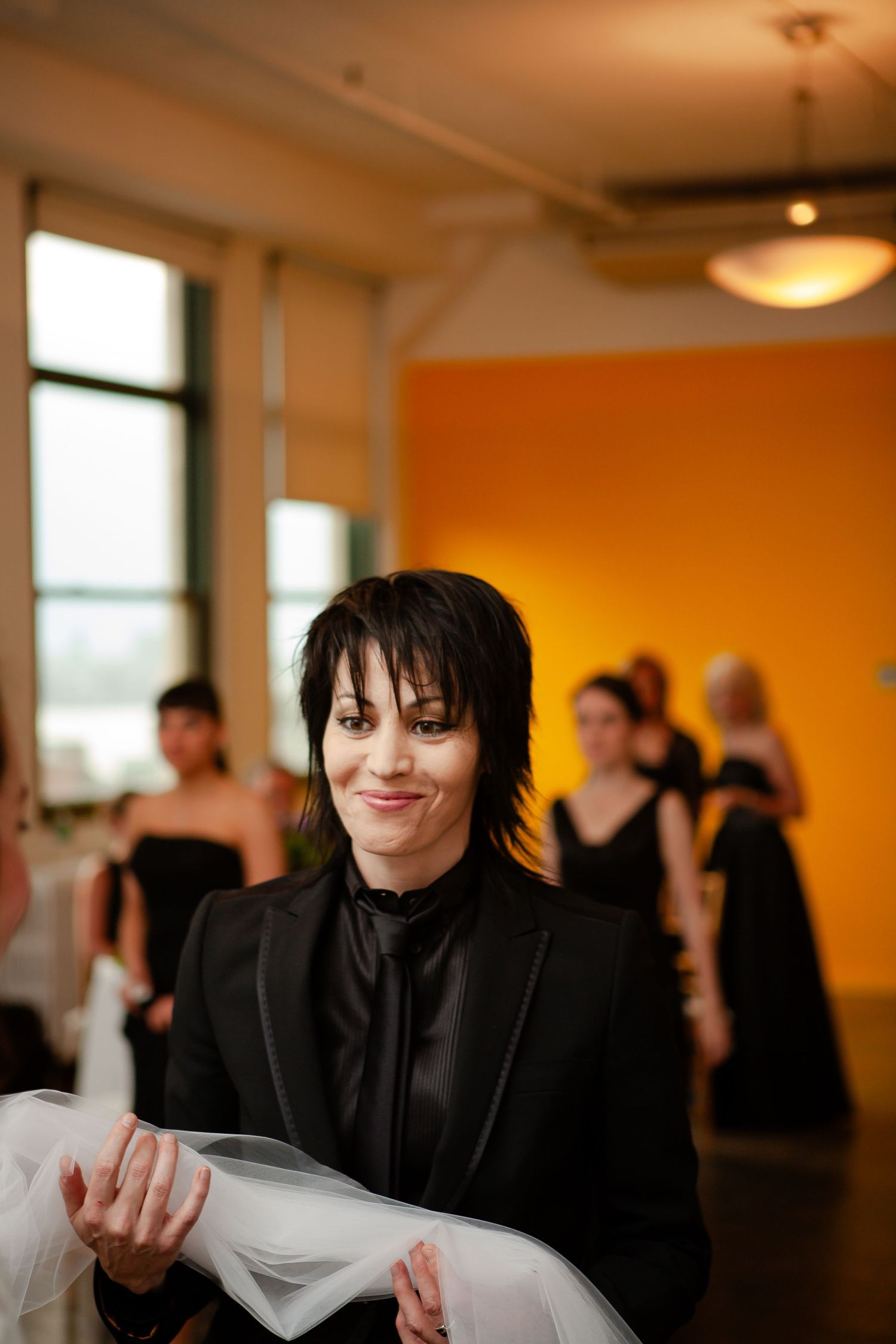 Person in black formal attire reading a document at an indoor event with orange walls and other guests in black dresses.