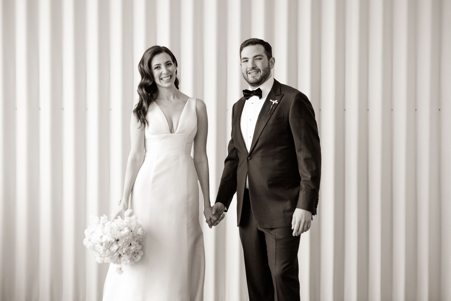 A black and white wedding photo of a couple holding hands against a striped background.