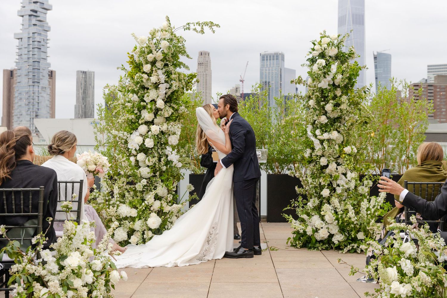 Wedding ceremony setup on rooftop terrace with white floral arch and city skyline view.