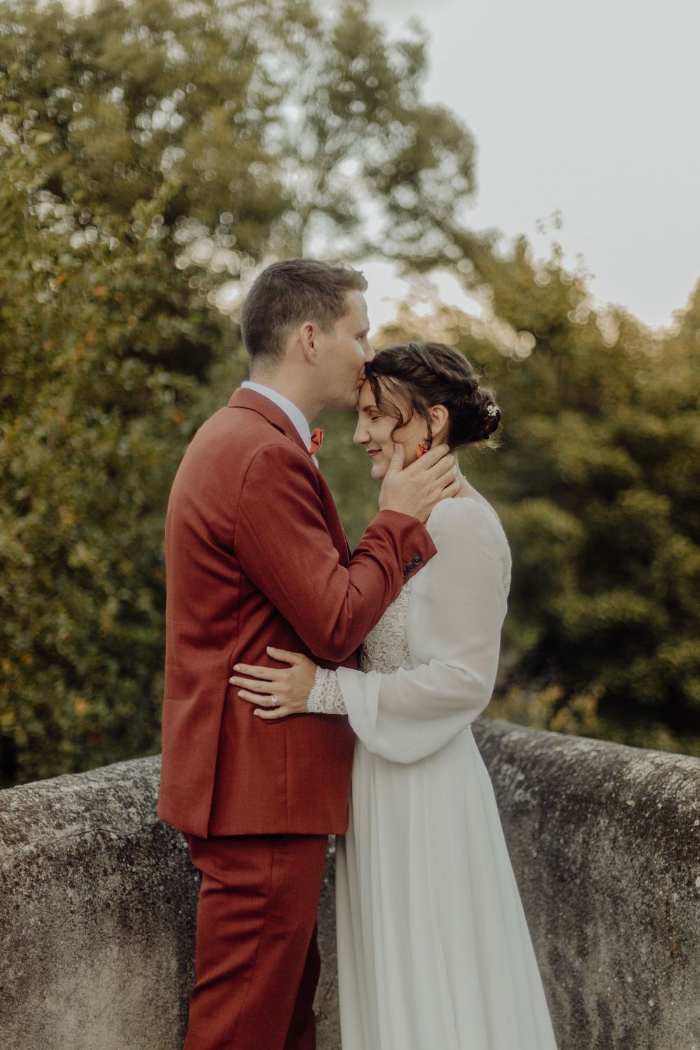 A couple in wedding attire share an intimate kiss against a stone wall and autumn foliage backdrop.