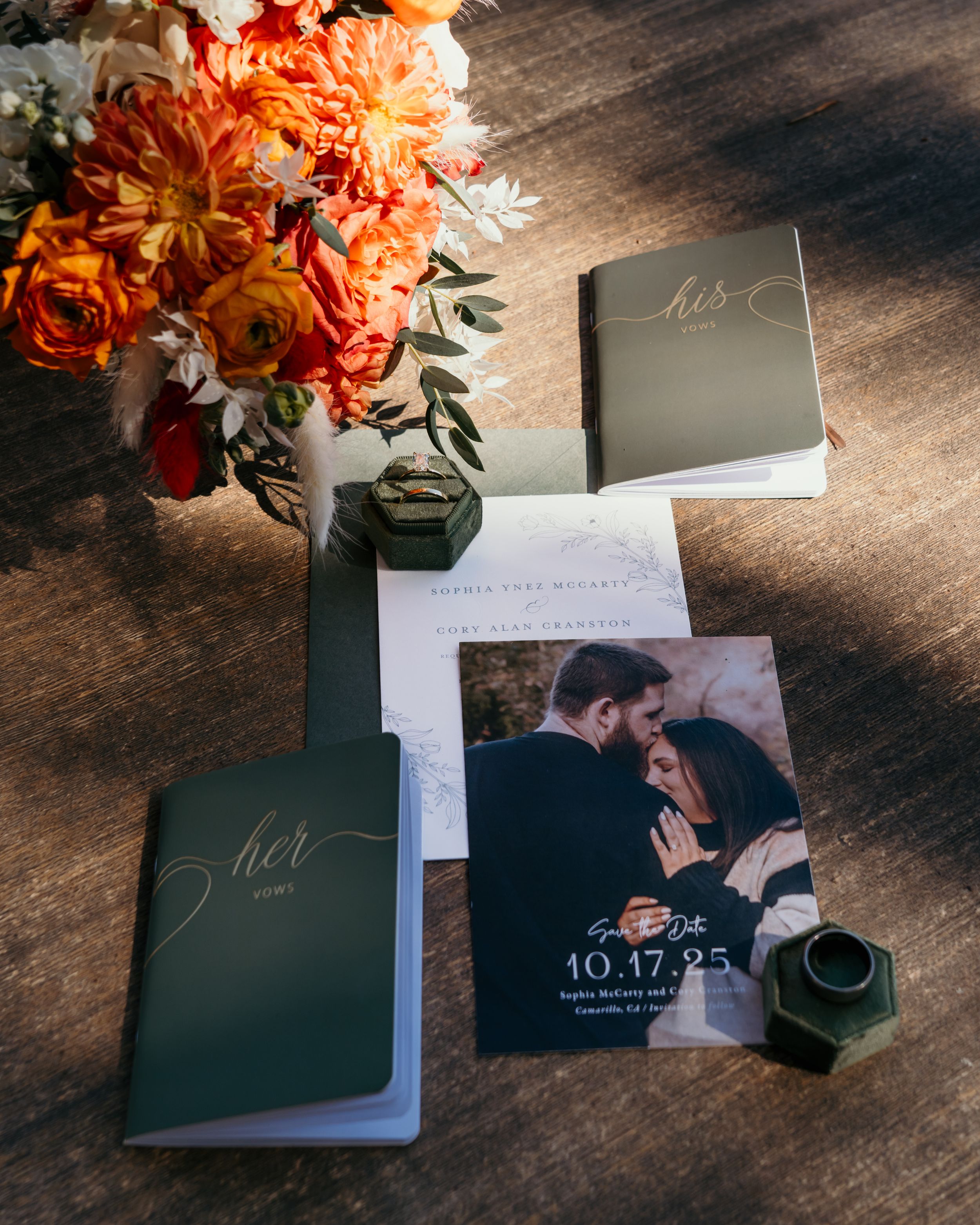 Wedding invitation suite with orange floral arrangements and engagement photos displayed on dark wooden surface.