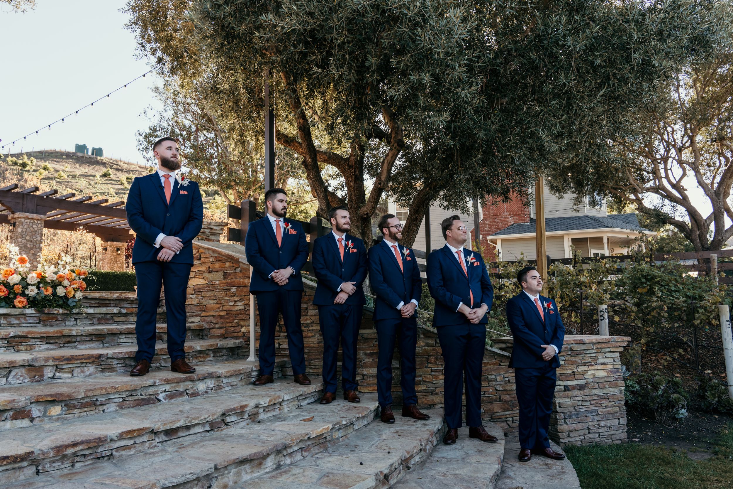 Wedding guests seated in wooden chairs at an outdoor ceremony venue with stone walls and trees.