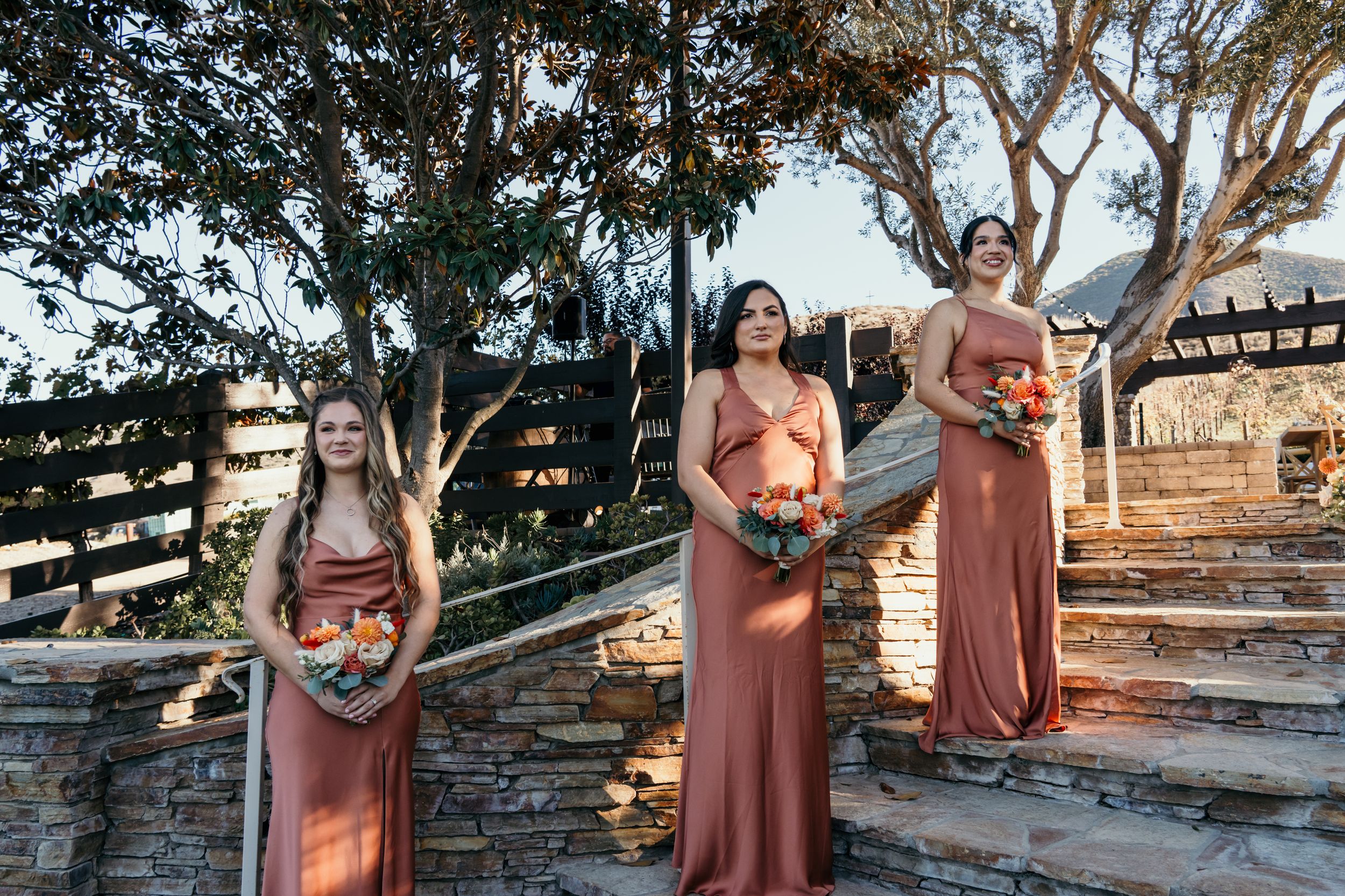 A group in rust-colored dresses poses together at sunset against a rustic stone wall and wooden fence backdrop.