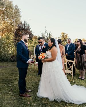 A wedding ceremony takes place outdoors on a grassy hillside with fall foliage in the background.