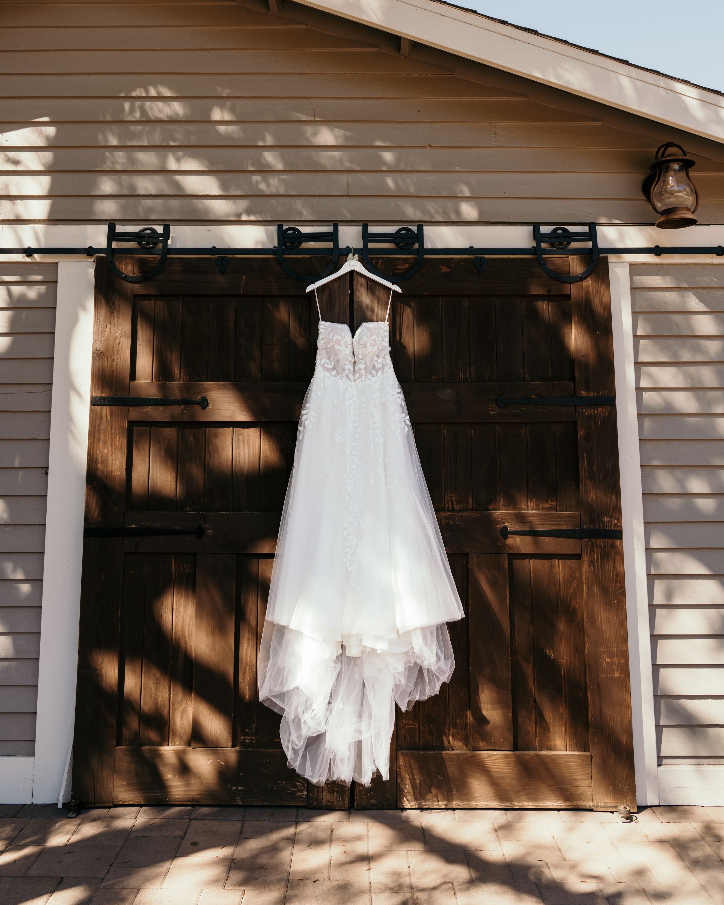A white wedding dress hangs elegantly against dark wooden barn doors with dappled sunlight creating beautiful shadows.