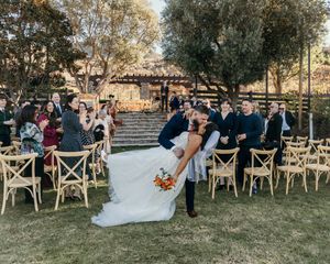 A panoramic view of an outdoor wedding ceremony setup with wooden chairs arranged on a lawn beneath trees at sunset.