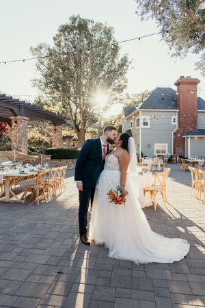 A romantic outdoor wedding reception setup with string lights, wooden tables and chairs on a stone patio at sunset.