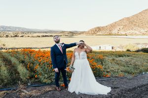 A couple stands together in a desert landscape with orange wildflowers and mountains in the background at sunset.