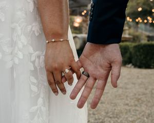 Close-up shot of newlyweds' hands with wedding rings and a delicate bracelet against white dress fabric.