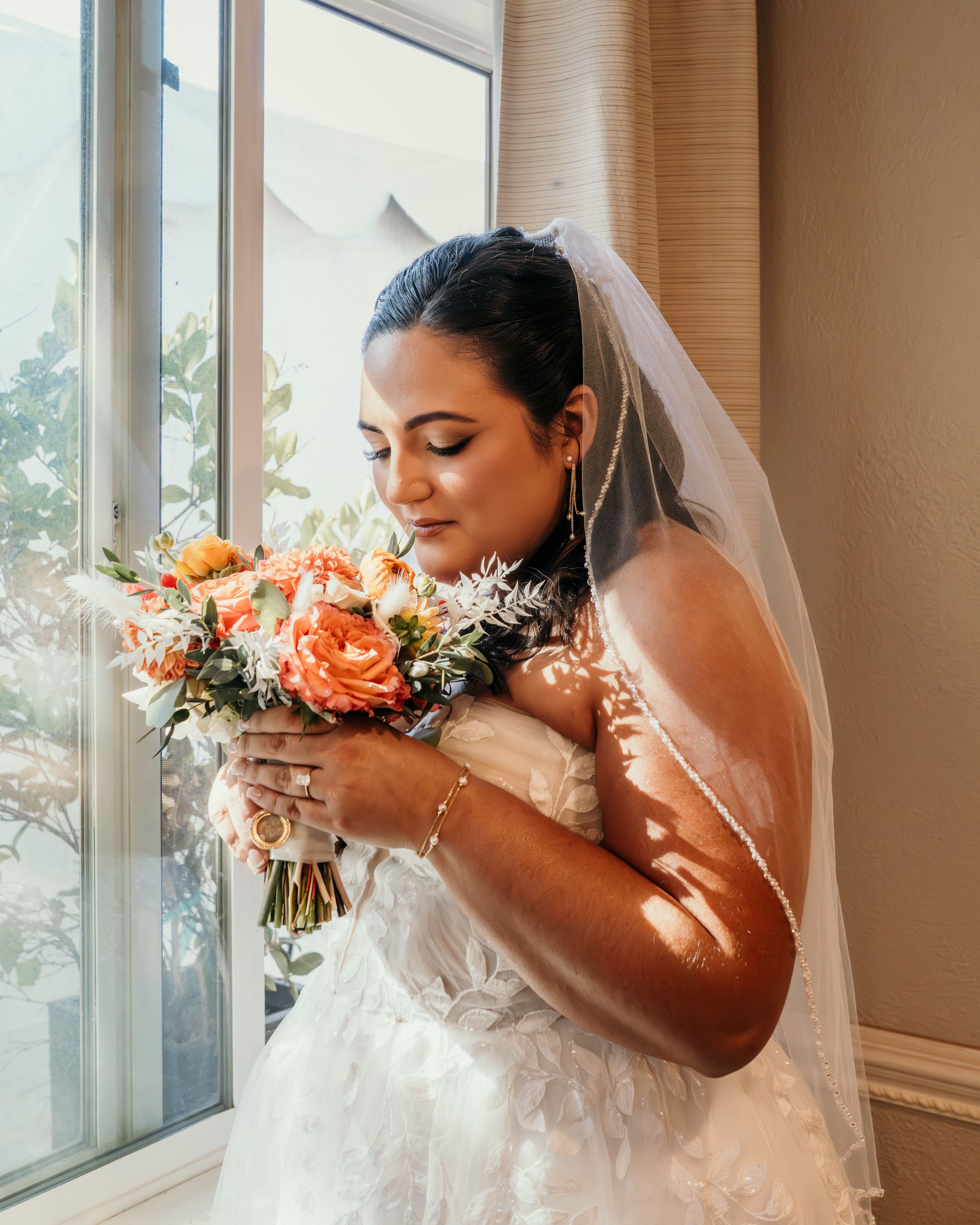 Bride in white dress holds and smells coral-colored wedding bouquet near sunlit window.