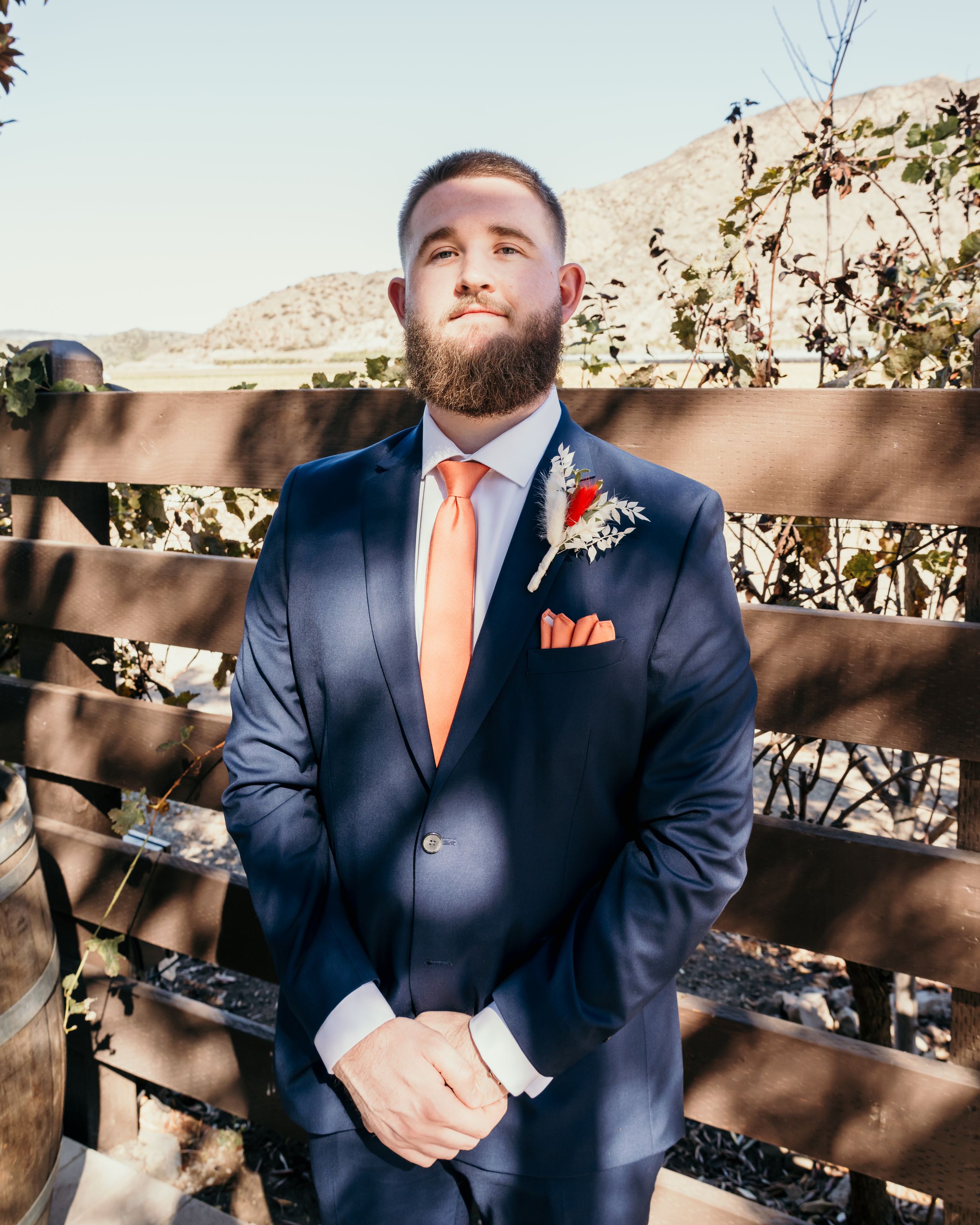 A groom in a navy suit with orange tie and pocket square stands in front of a rustic wooden fence.