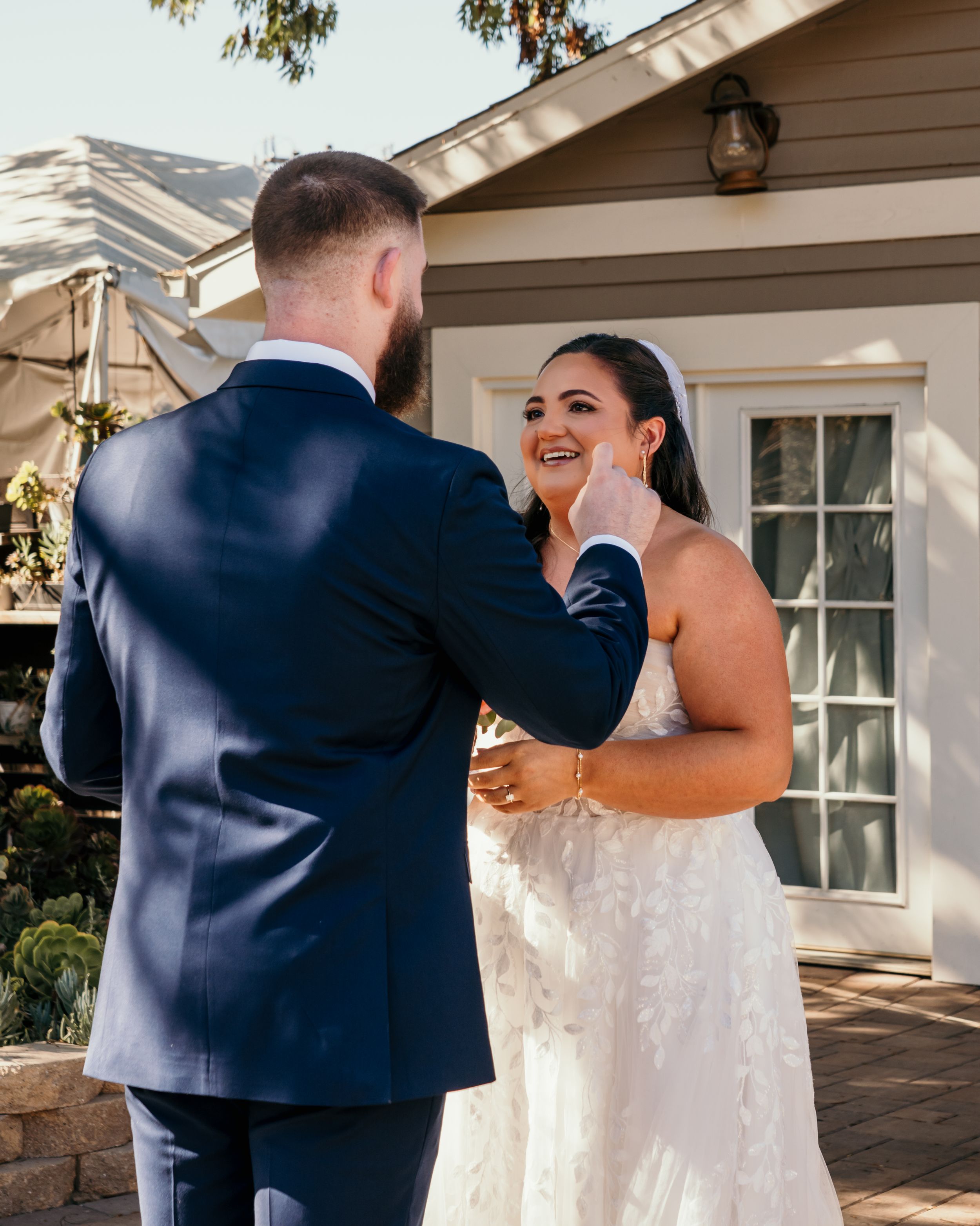 A joyful moment between a bride in a white lace gown and groom in navy suit during their first look outside a house.