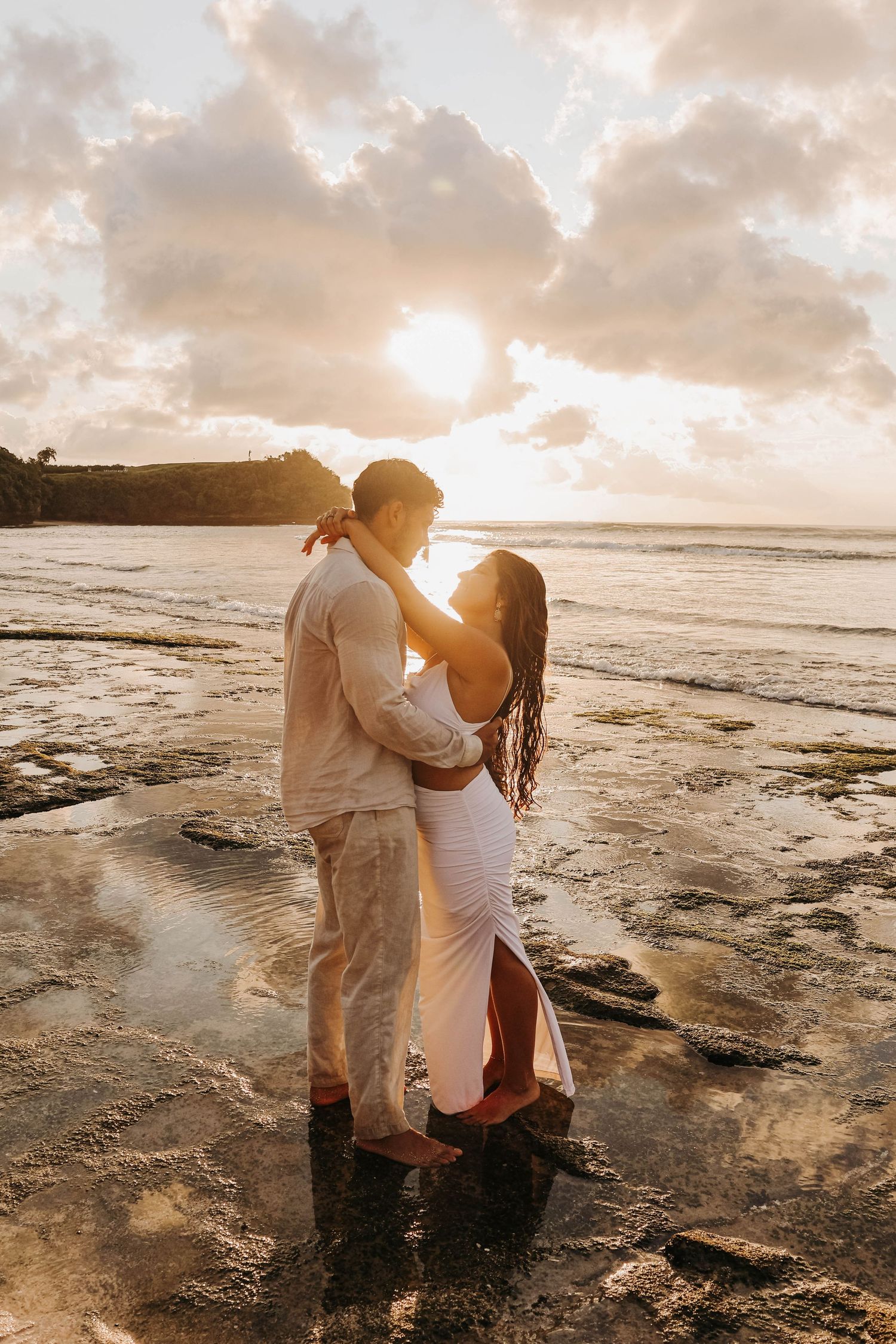 Happy couple in white outfits sitting on sandy beach, woman laughing joyfully while man embraces her from behind.