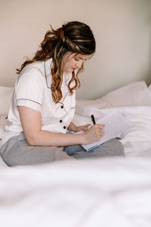 A woman in a white shirt writes in a journal while sitting on a bed with white linens.