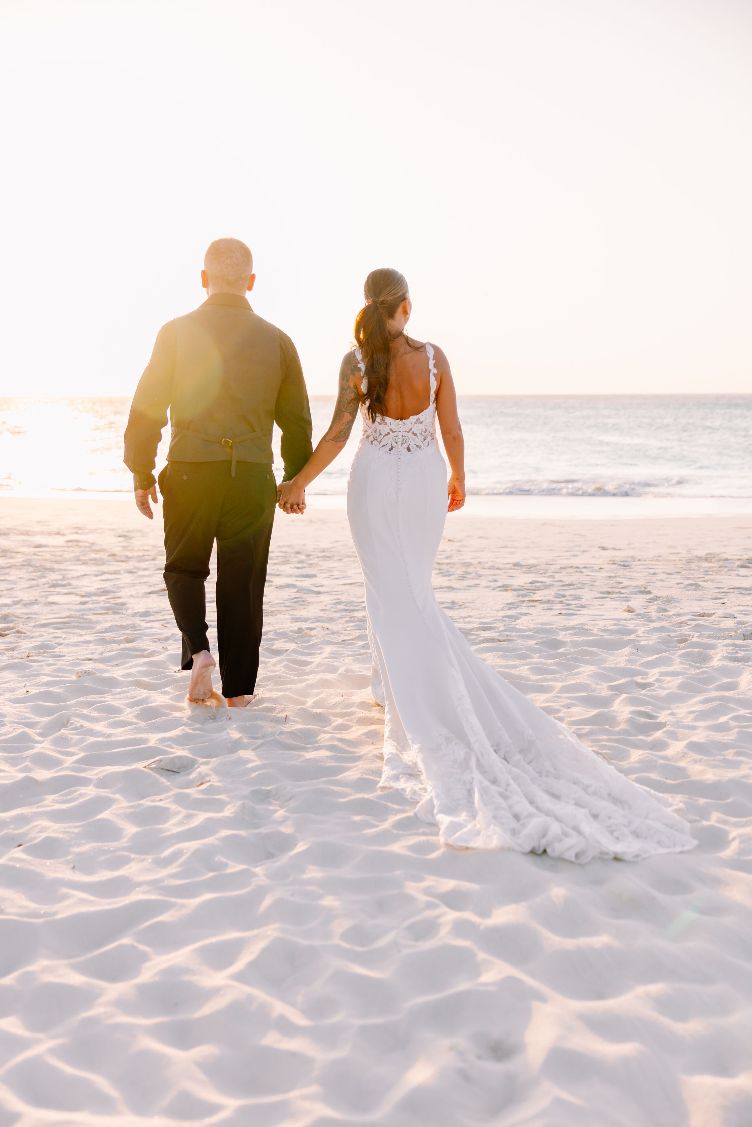 Couple walking together on a white sandy beach at sunset with the bride's flowing white dress trailing behind them.