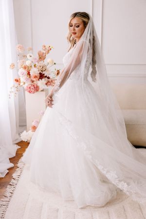A stunning bridal portrait featuring a flowing white wedding gown and veil against a light backdrop with pink floral accents.