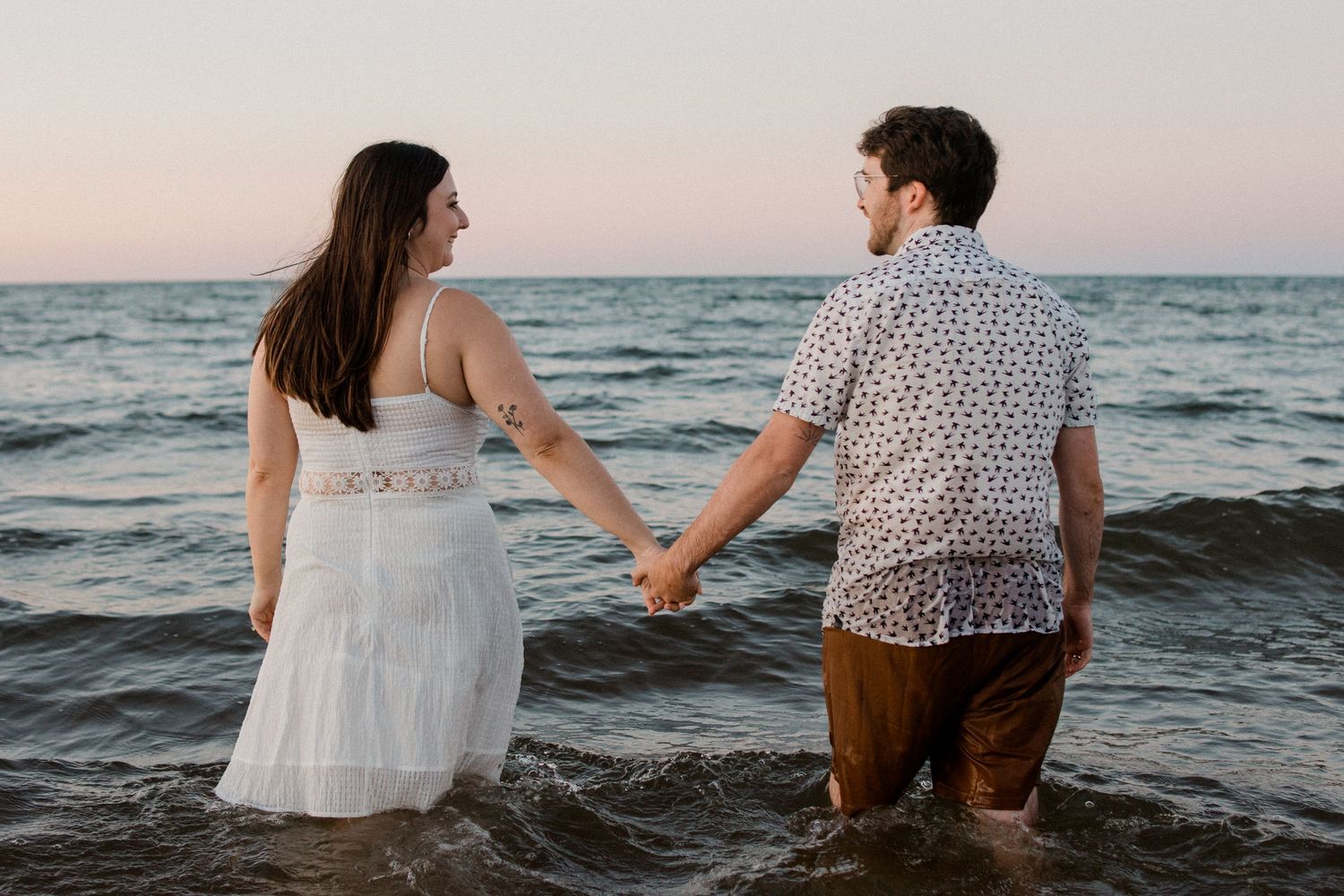A couple holds hands while standing in shallow ocean waves at sunset, with the person on the left wearing a white dress.