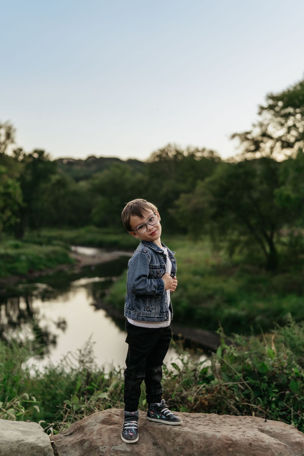 A small child in denim clothing stands on rocks near a serene river during golden hour for family photo session in Akron, Ohio's Metro Park