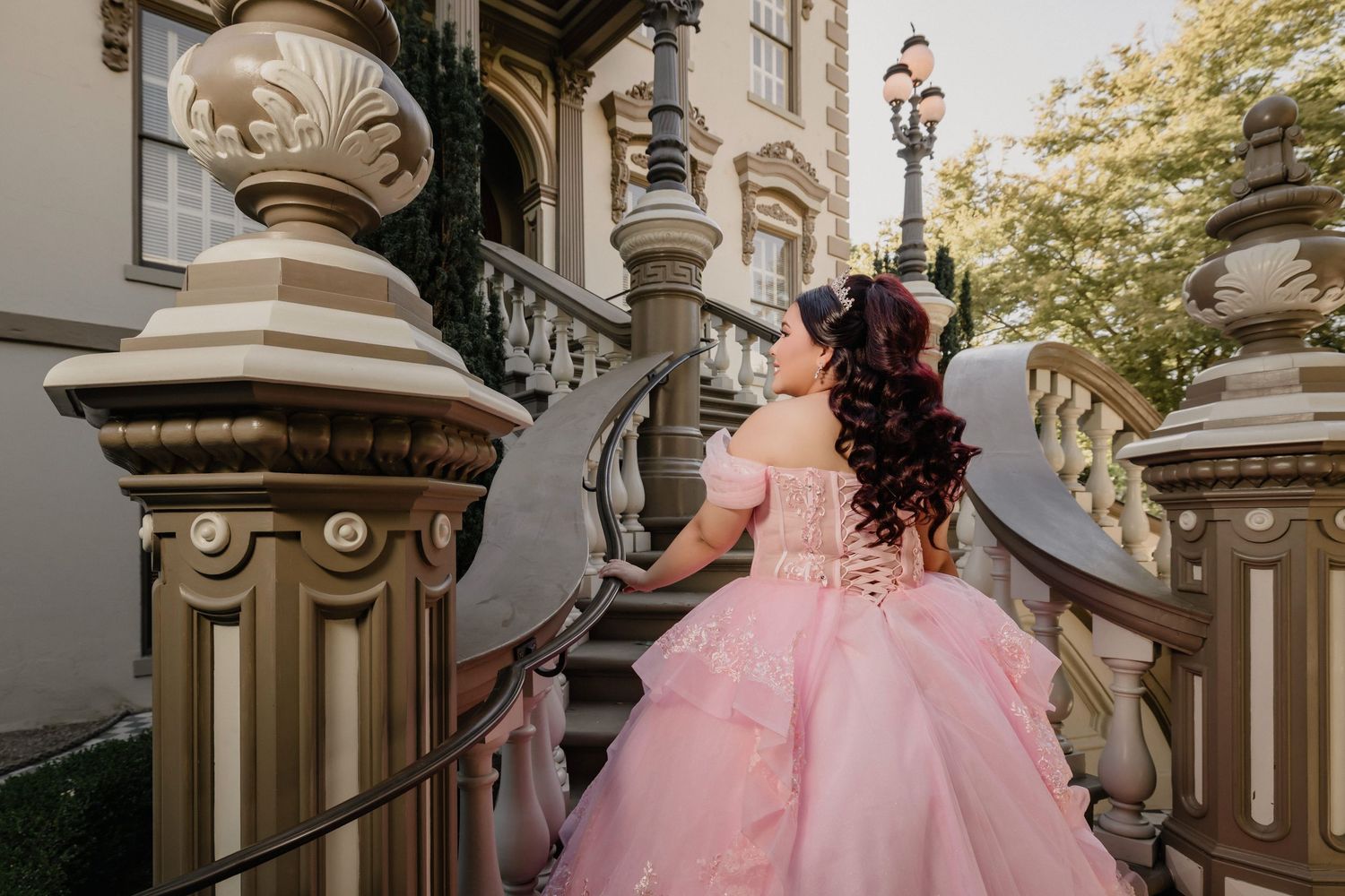 A person in a pink ballgown poses on elegant white stone steps with ornate architectural details.