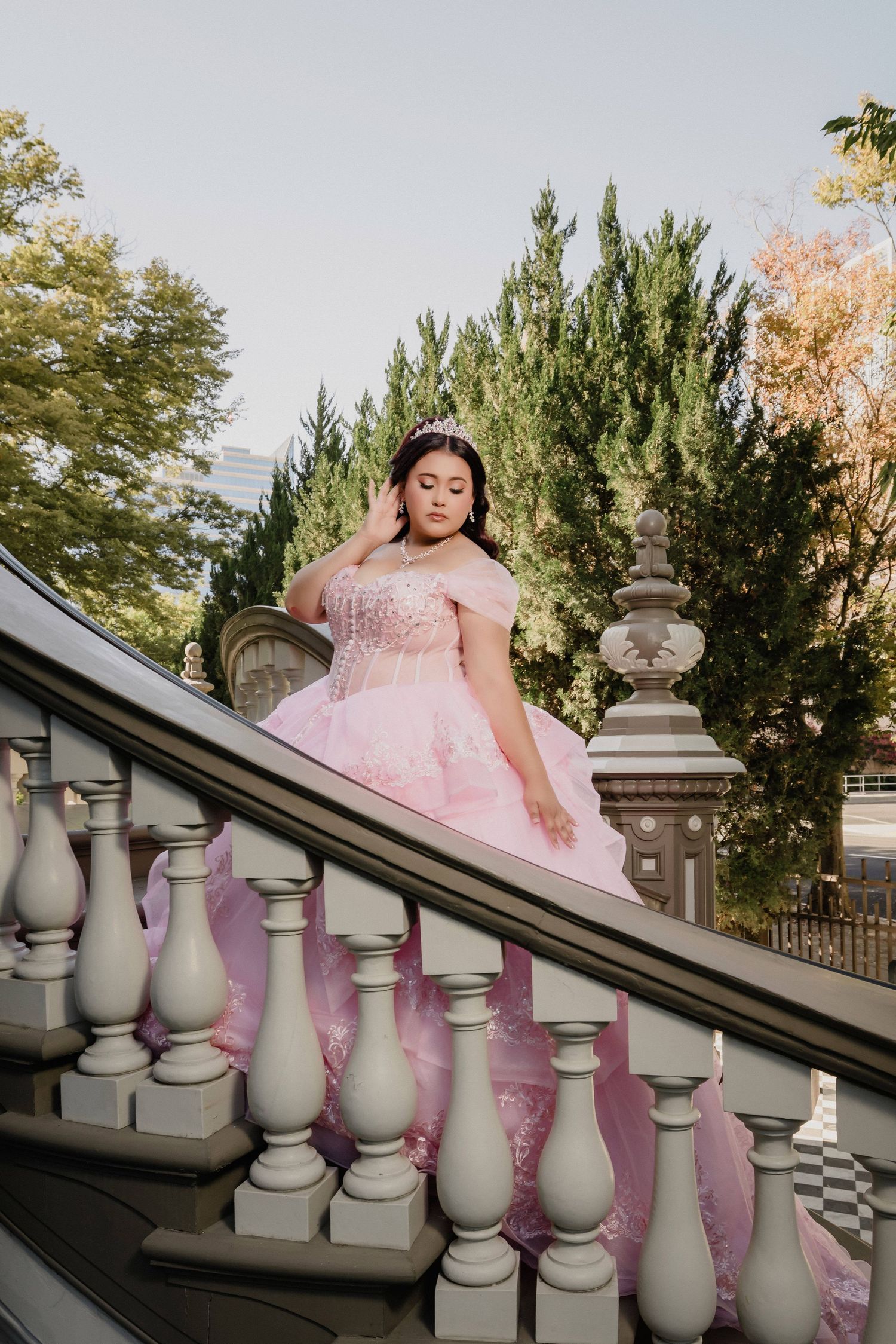 Pink princess gown with intricate embroidery photographed against white stone balustrade and evergreen trees.