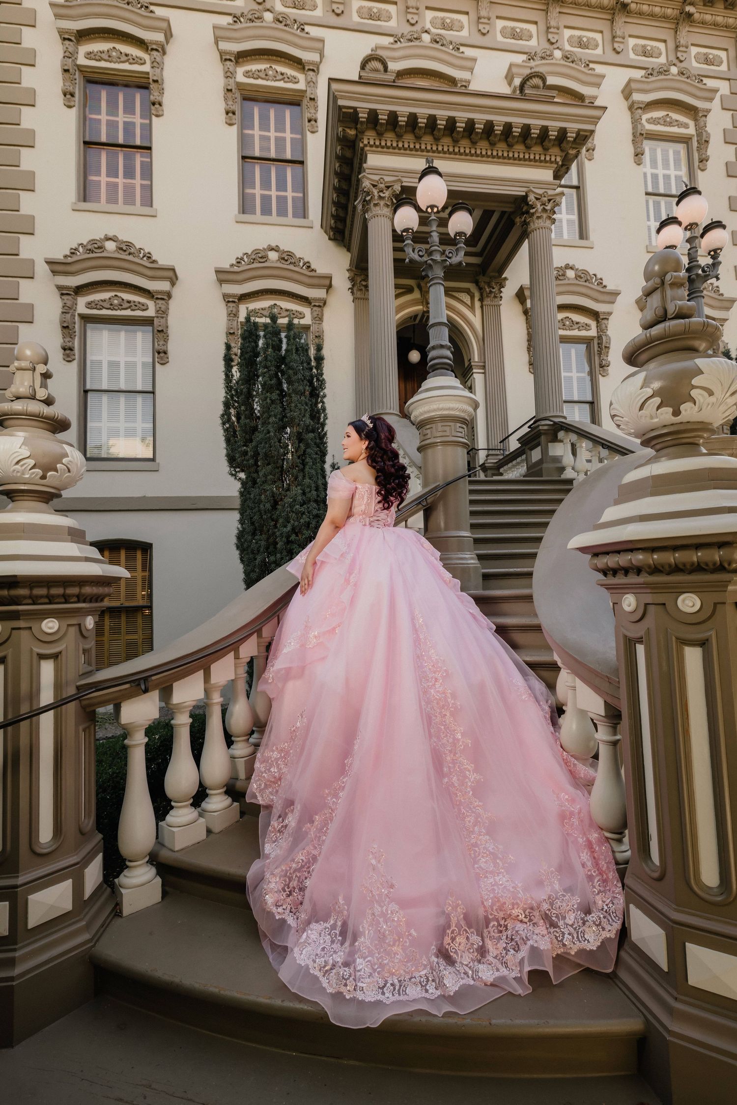 Elegant pink quinceanera gown with lace details shown against classic architectural backdrop.