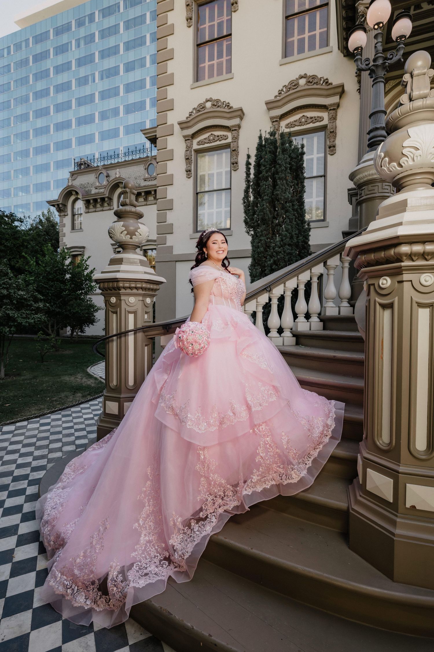 Elegant pink ballgown with lace details photographed on grand marble staircase with checkered floor pattern.