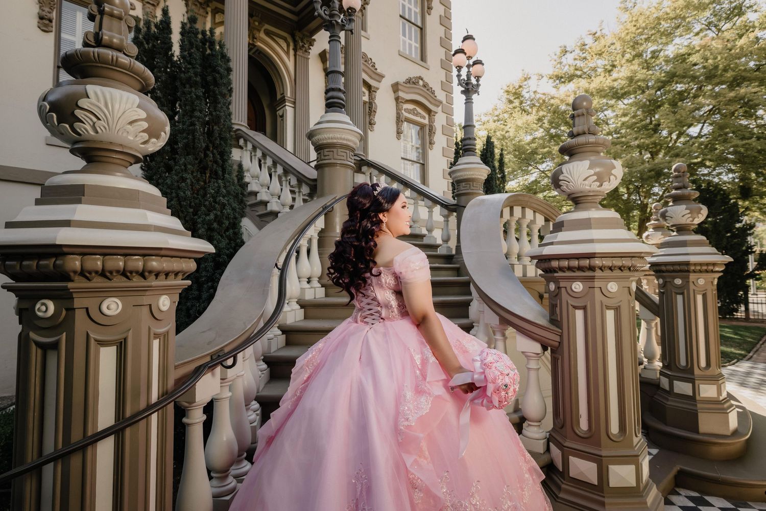 Elegant pink ballgown photographed on a grand staircase with ornate architectural details and checkered floor.