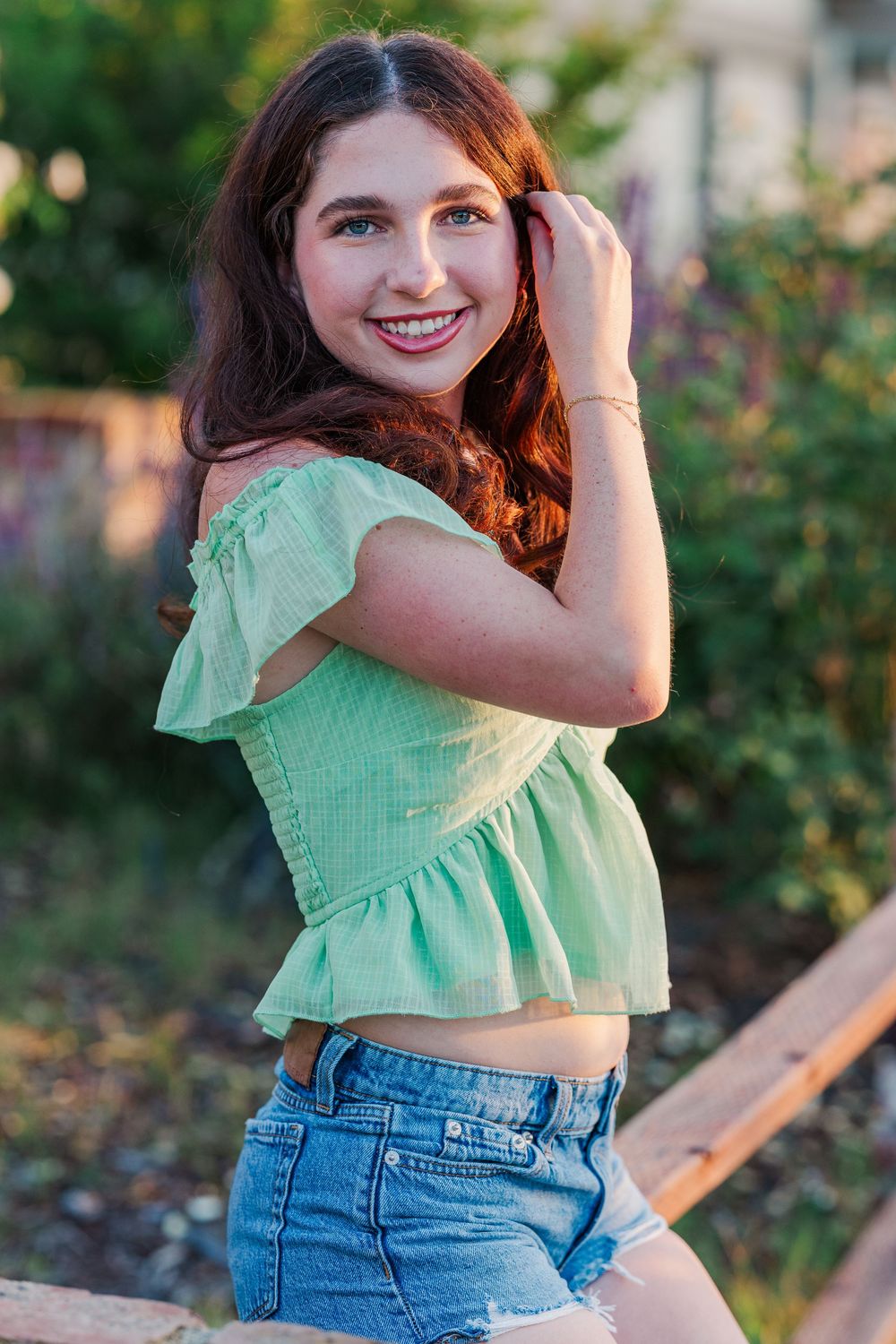 A person in a mint green crop top and blue jeans poses playfully in a garden setting at sunset.