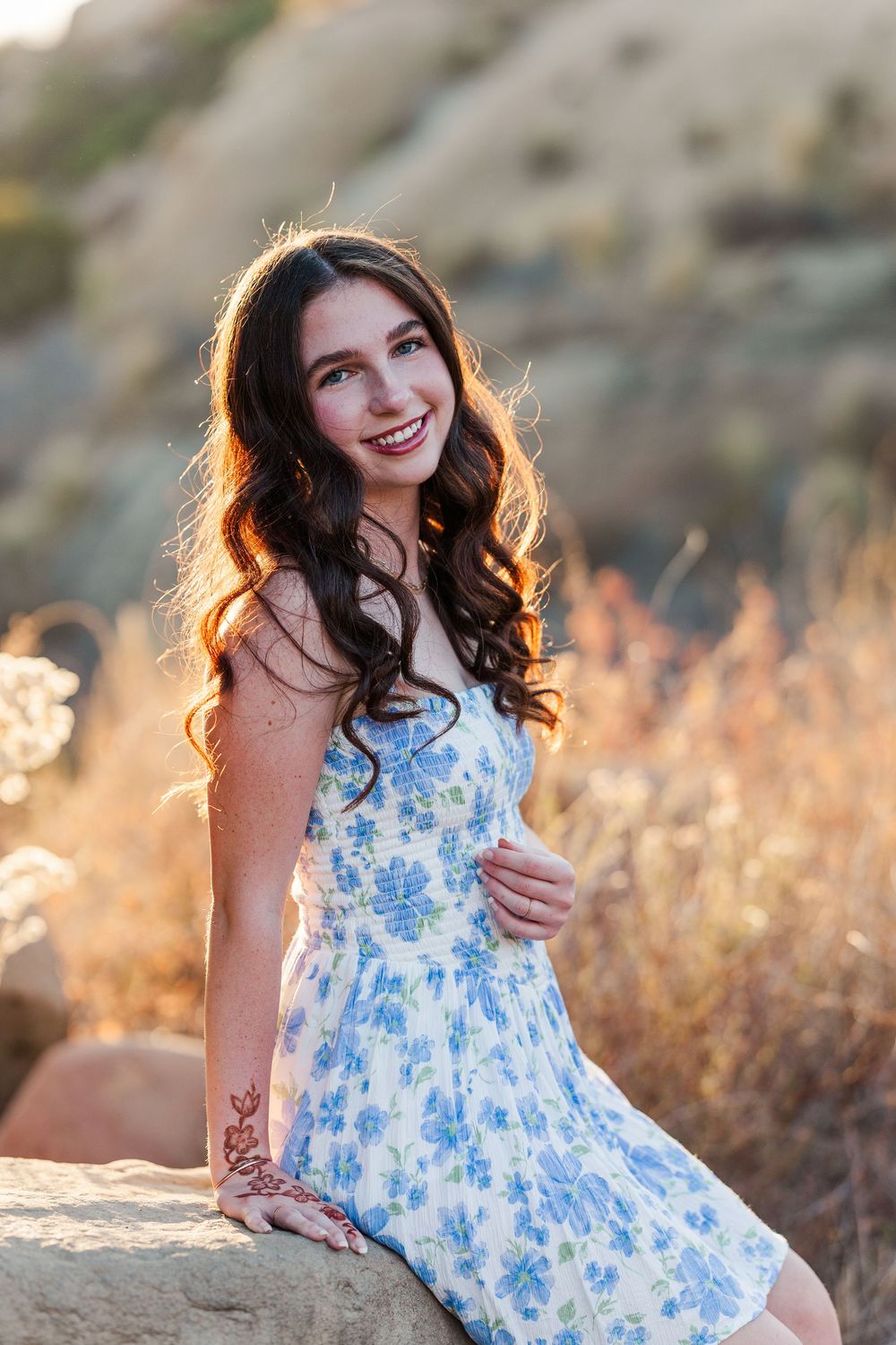 A person wearing a blue floral sundress sits on rocks during sunset with warm lighting.