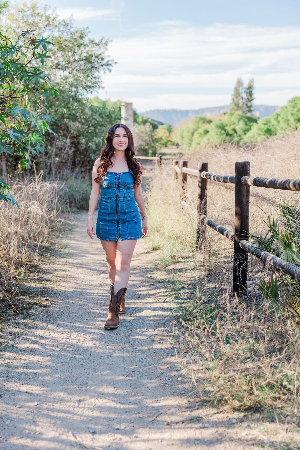 A sunny outdoor portrait on a dirt path with wooden fencing and mountains in the background.