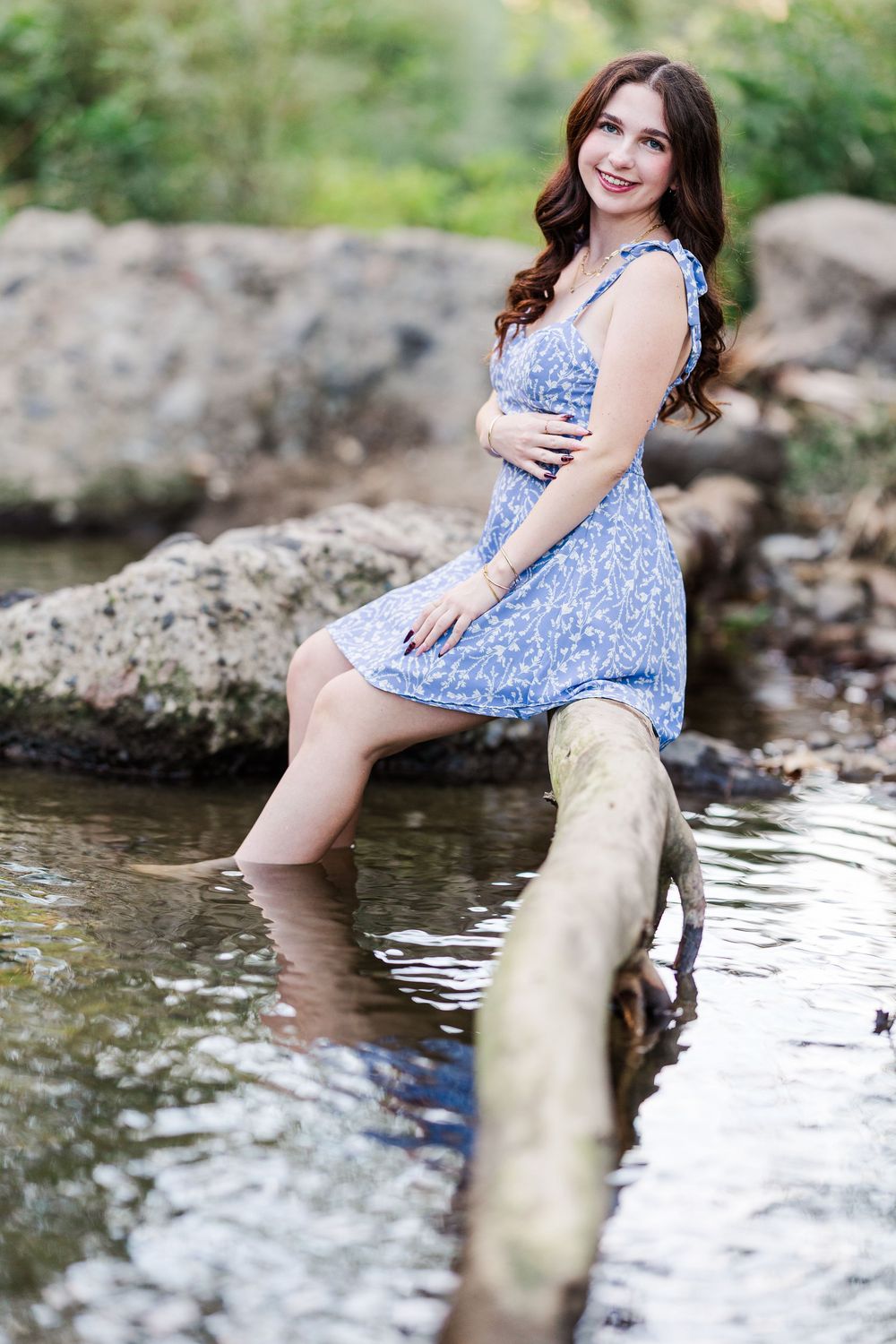A person in a blue floral dress poses playfully on a log by a stream in a summer nature setting.