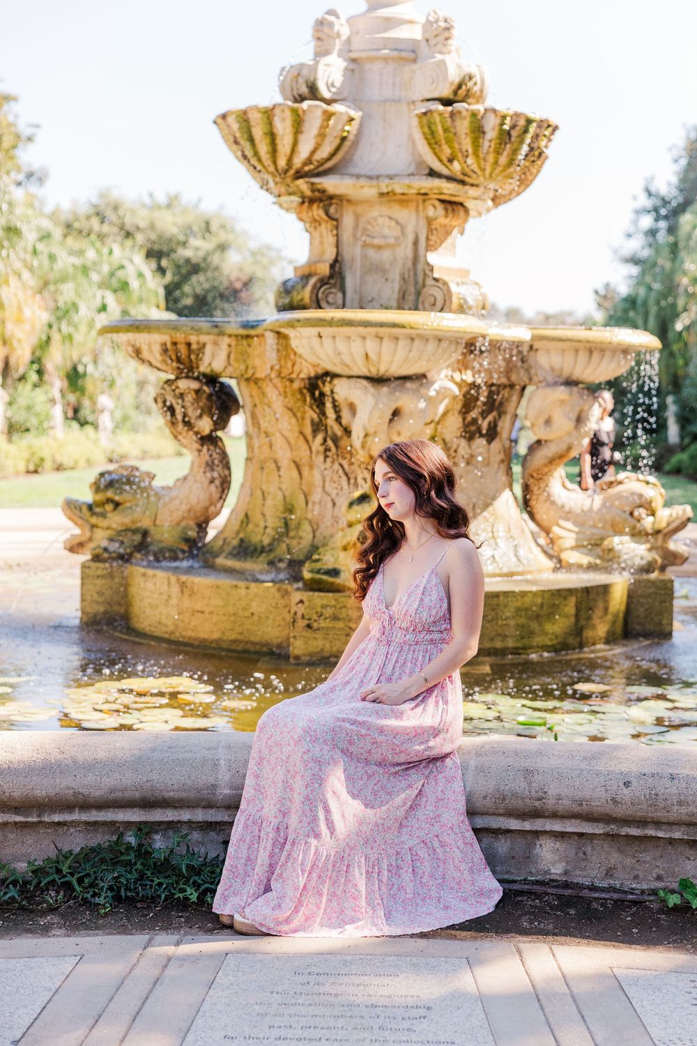 A person in a flowing pink dress poses gracefully by an ornate stone fountain in a garden setting.