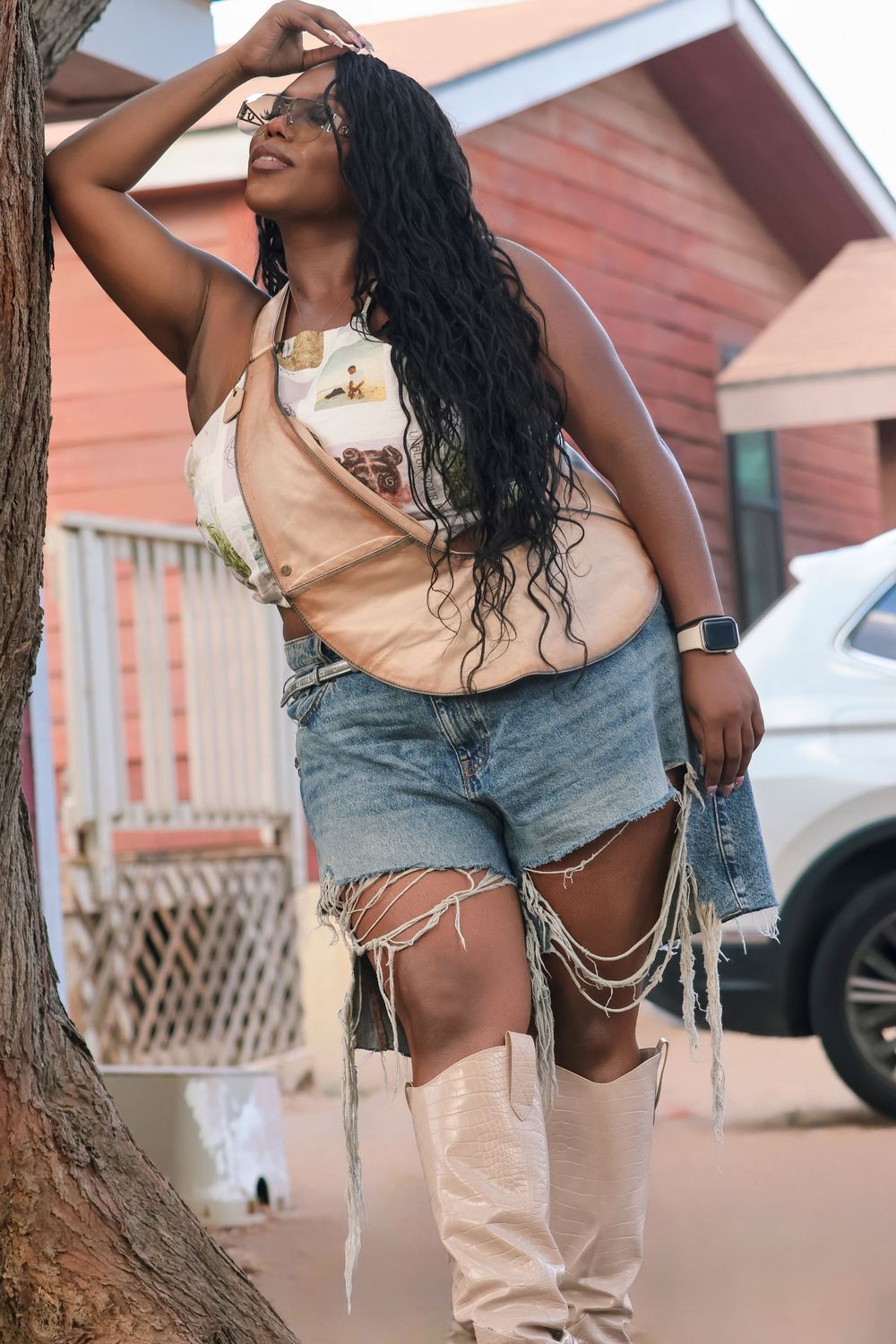 Woman in denim shorts and beige top posing casually against rustic backdrop.