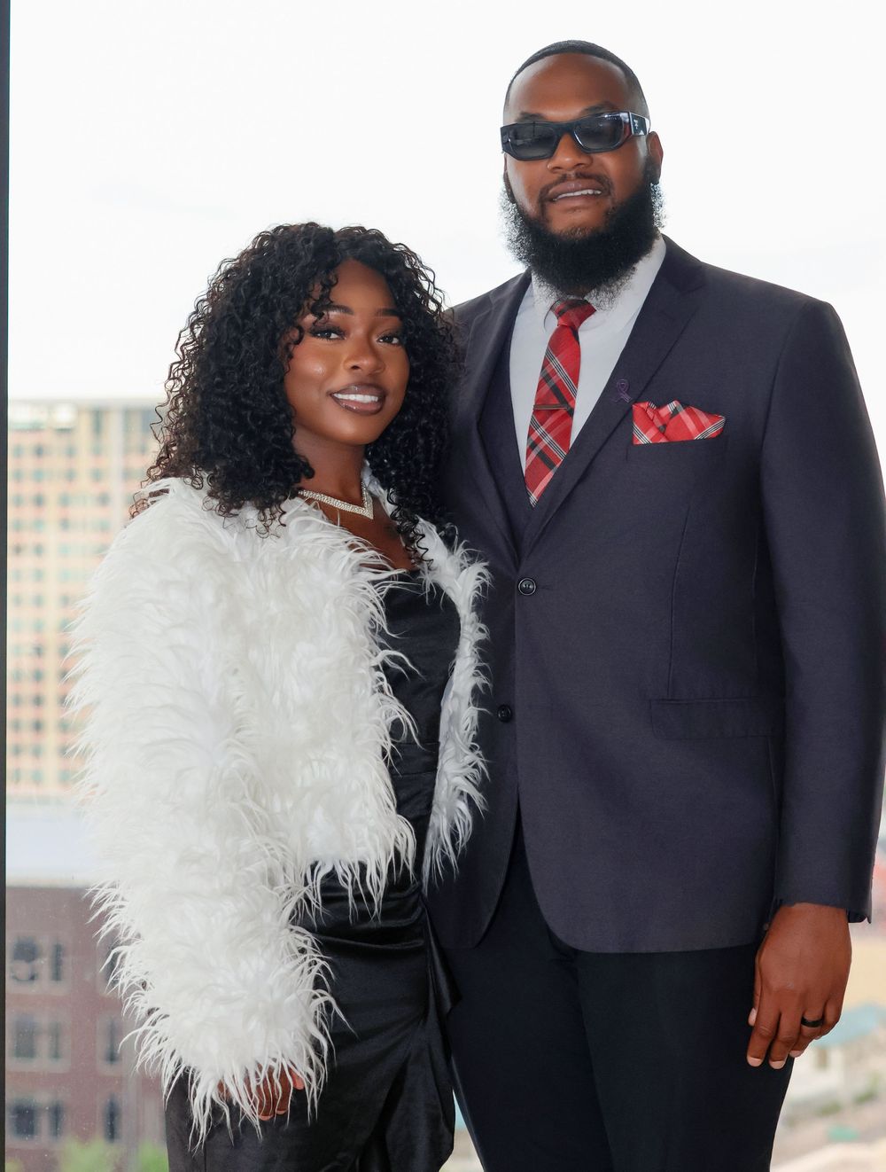 Couple posing together, with one wearing a white feathered accessory and the other in a dark suit.