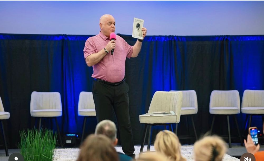A speaker in a pink shirt holds up a device while presenting on stage at a conference with white chairs in the background.