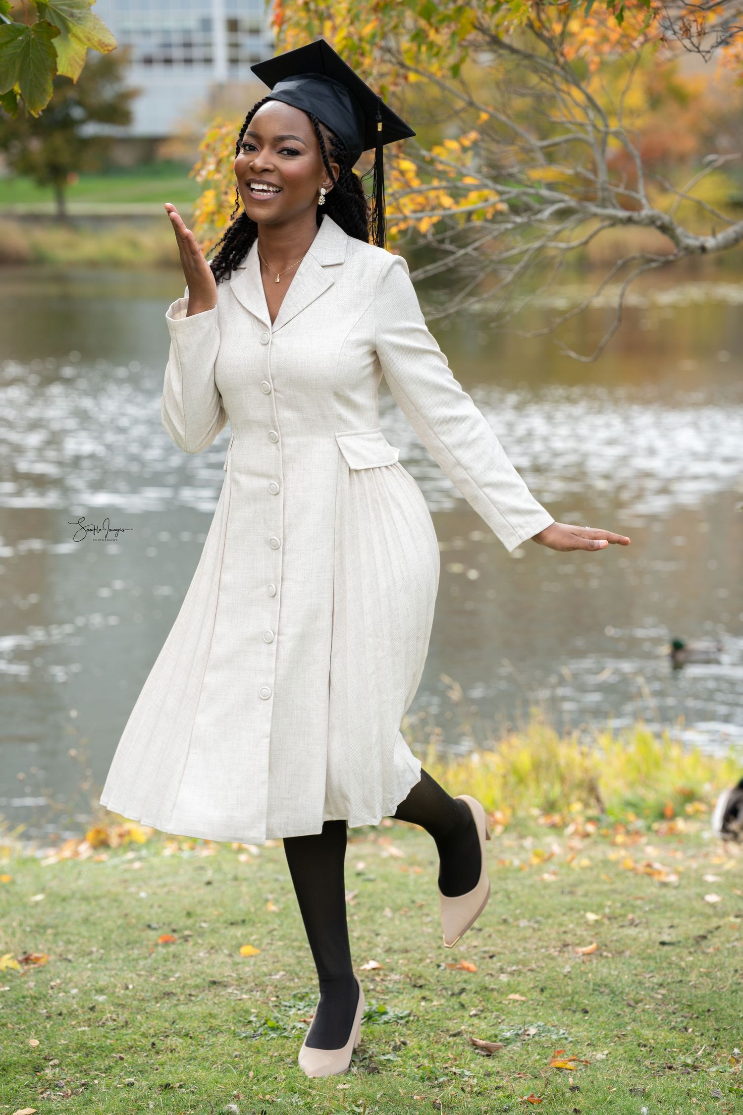 Graduate in white coat and cap poses joyfully by autumn lake with golden leaves in background.