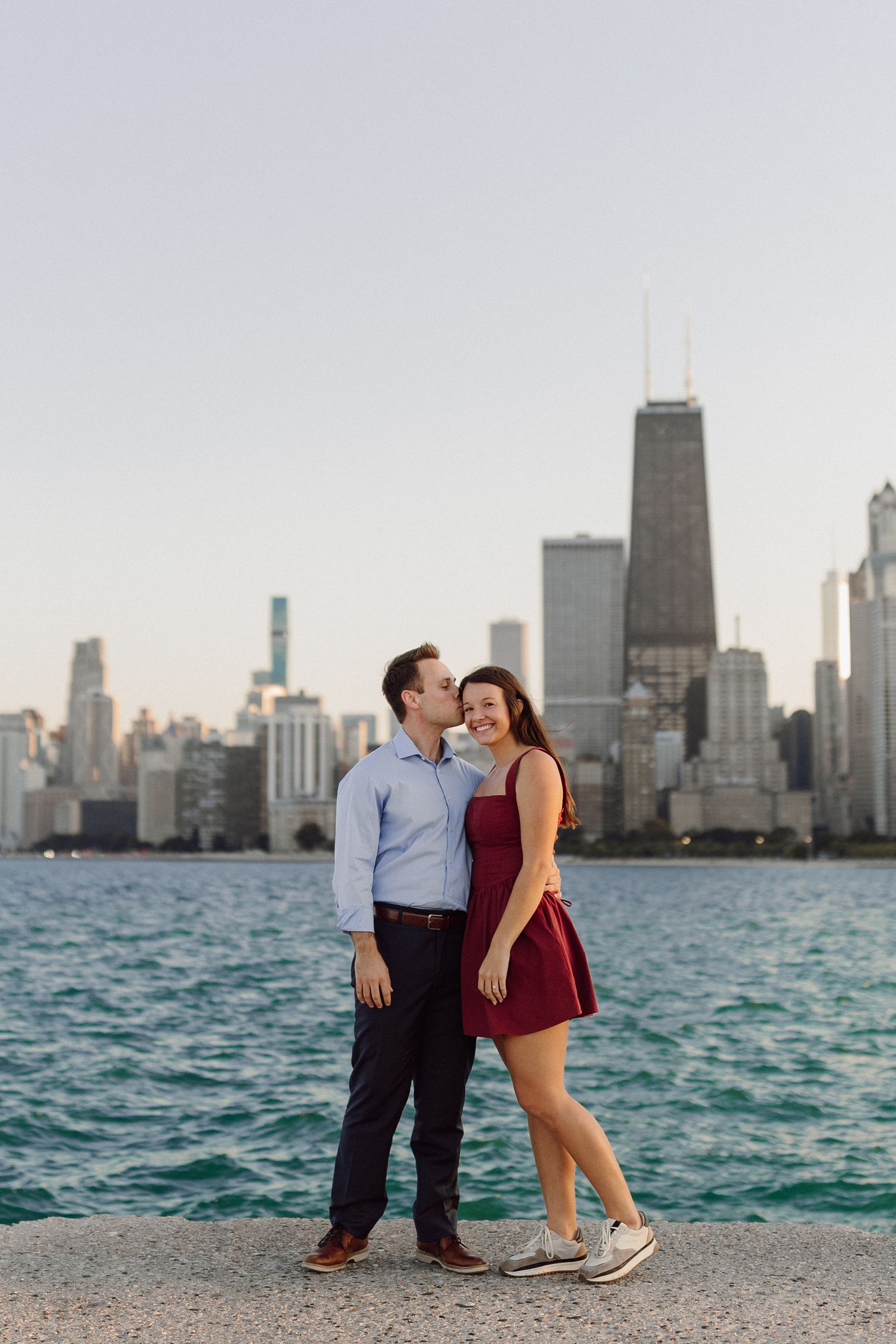 Couple posing by Lake Michigan with Chicago skyline in background during sunset.