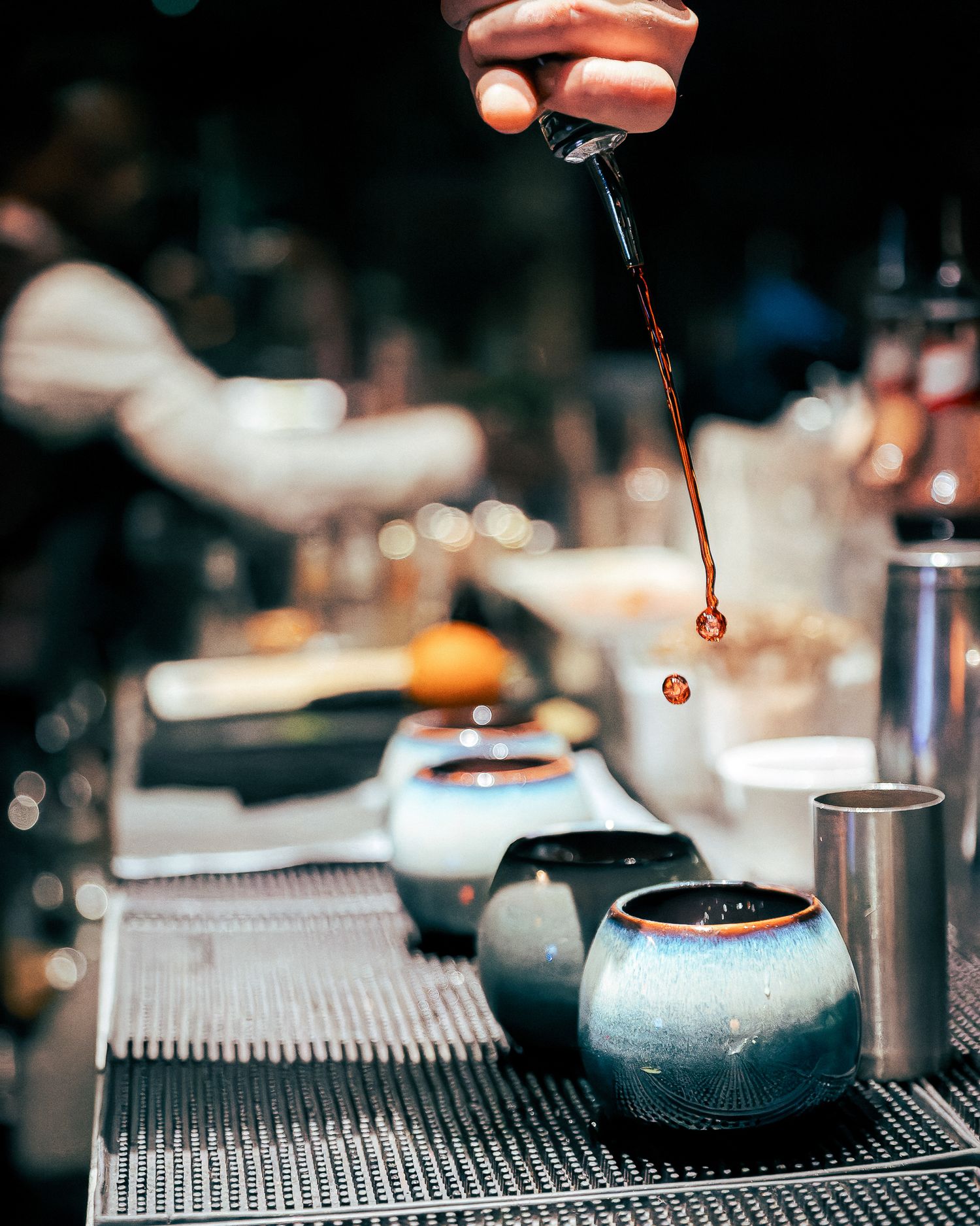 Bartender pouring drinks into ceramic vessels on a metallic bar surface.