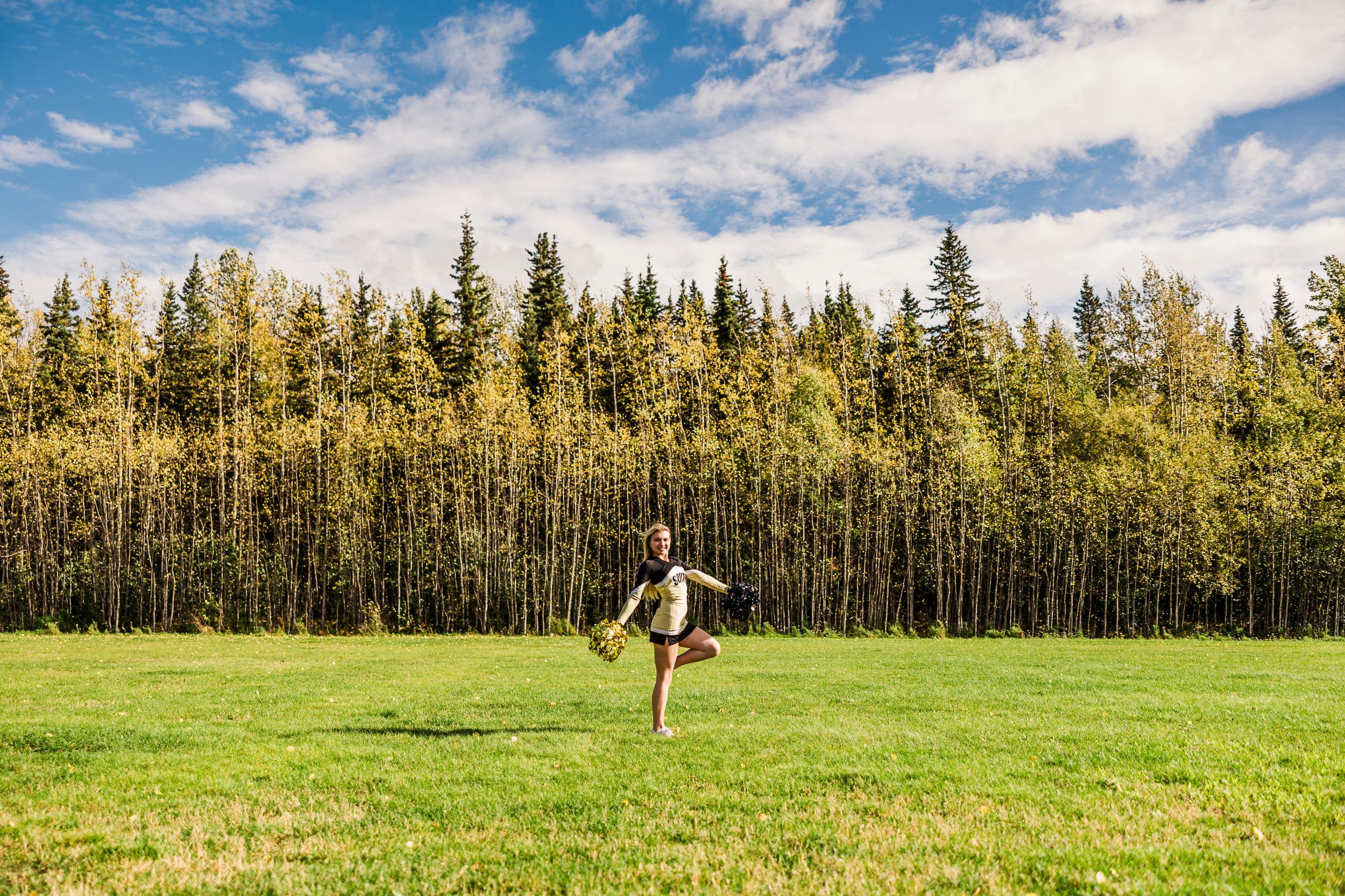 A person in athletic wear poses on a turf field with yellow and black pom poms for a sports-themed photo session.