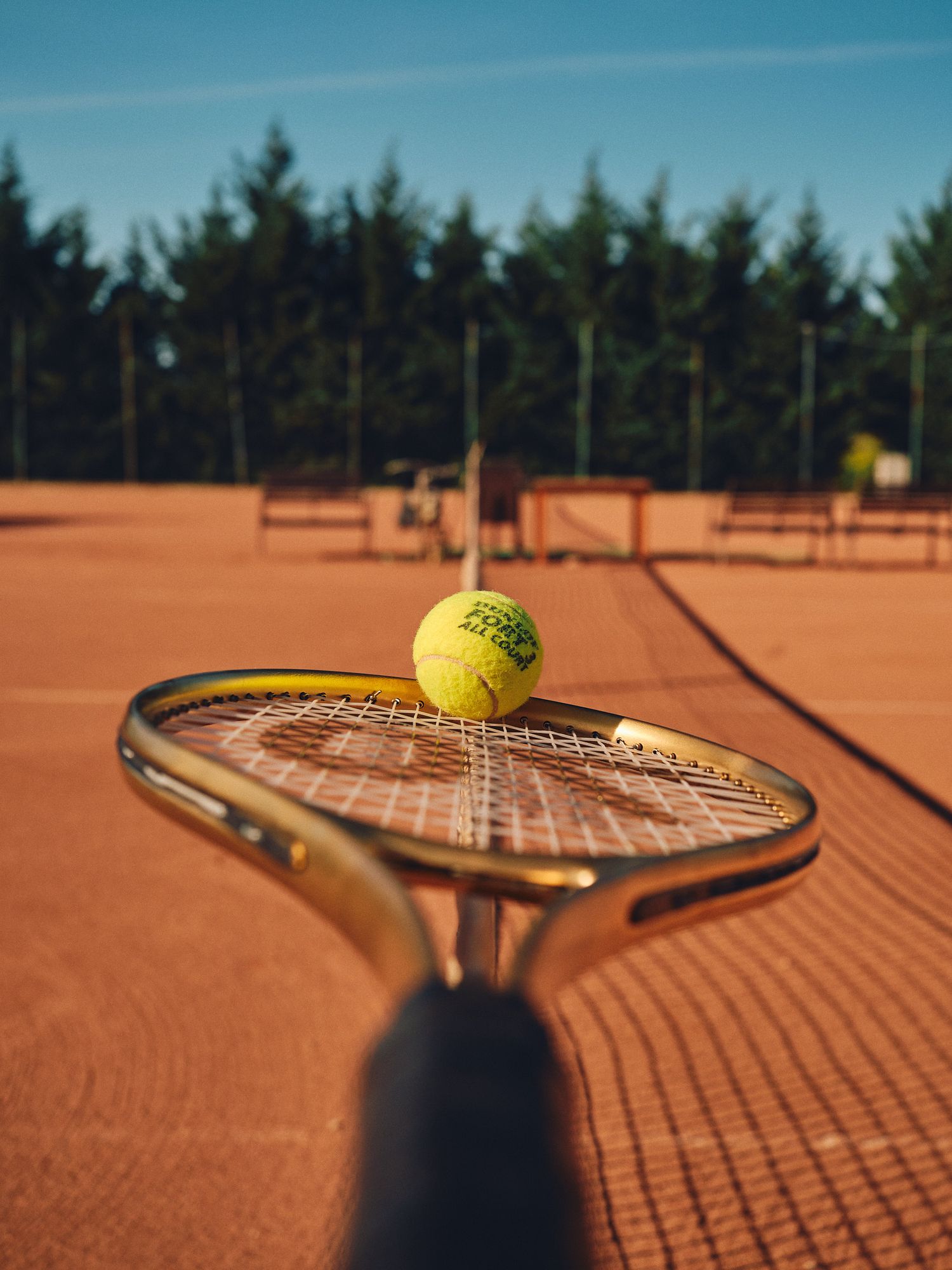 Tennis ball and racket resting on a clay court with shadows casting dramatic angles in warm sunlight.