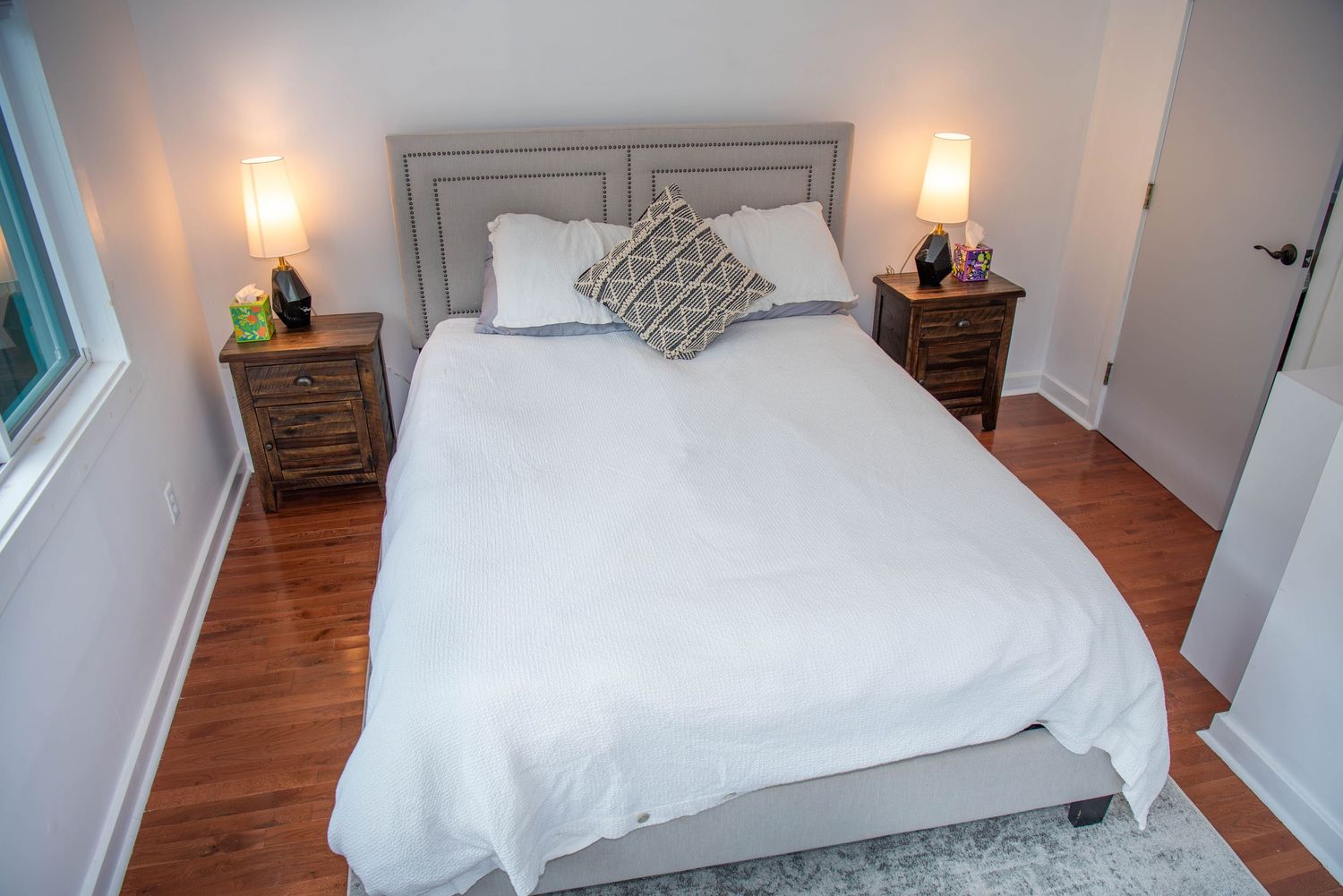 Modern bedroom interior with white bedding, hardwood floors, and natural light from windows.