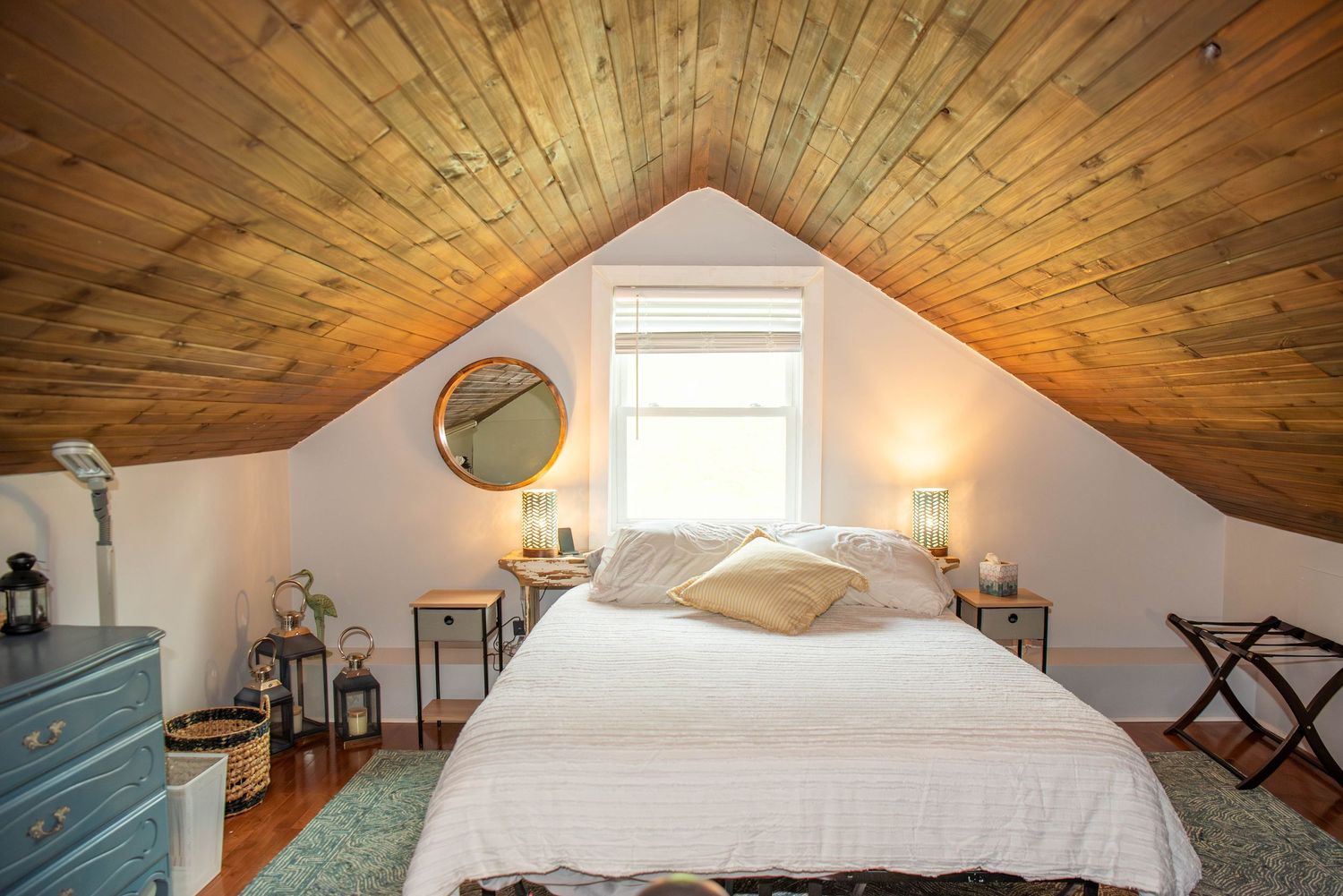 Cozy attic bedroom with wooden vaulted ceiling, white bedding, and natural light streaming through a window.